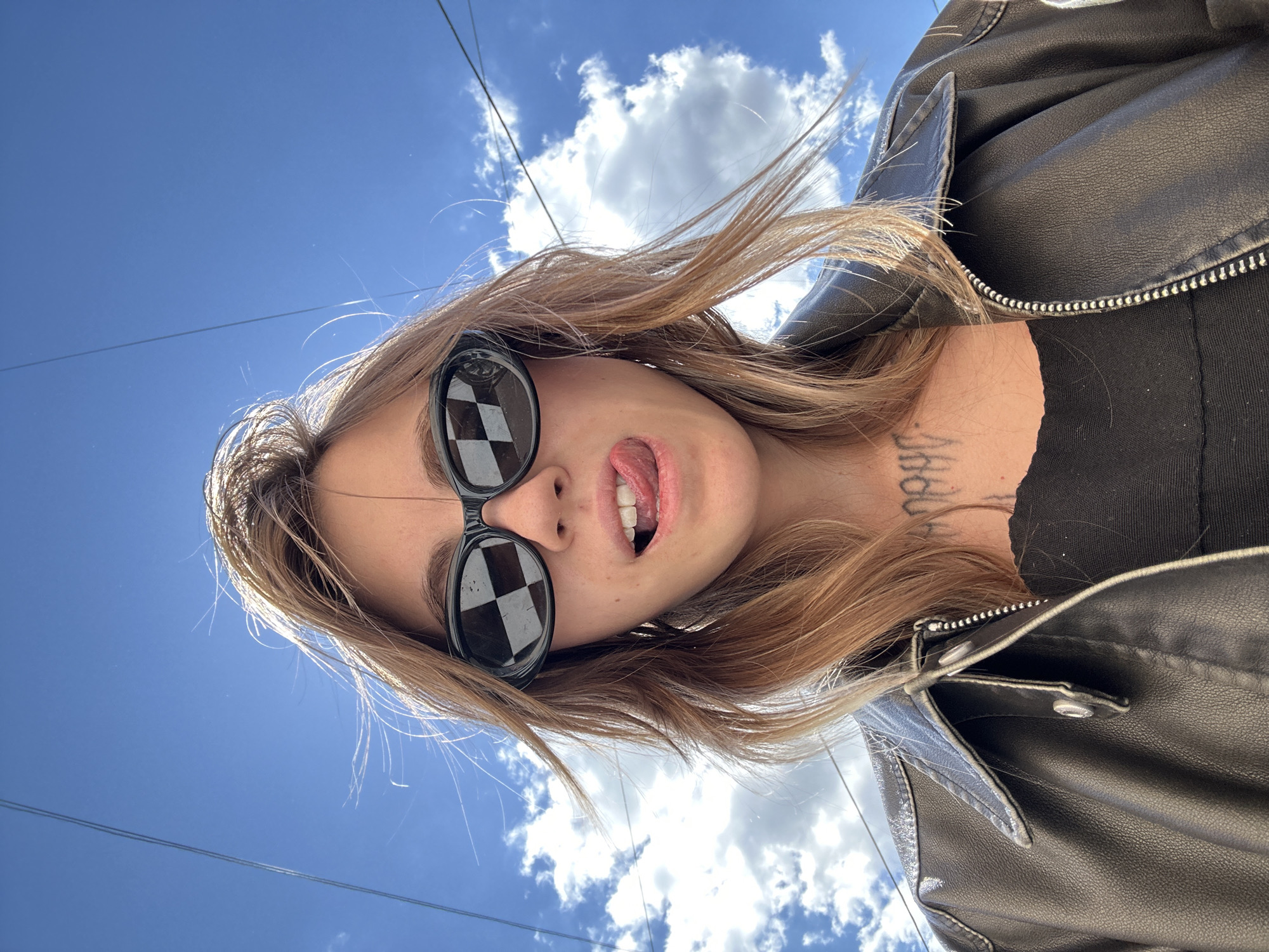 A woman with blonde hair and sunglasses sticks her tongue out at the camera. She is wearing a black leather jacket and a black top. The background is a bright blue sky with white clouds and some power lines. There is a tattoo on her neck. The image appears to be taken outdoors during the daytime in a location that could be Moscow, Russia, given the presence of power lines and the general urban aesthetic.