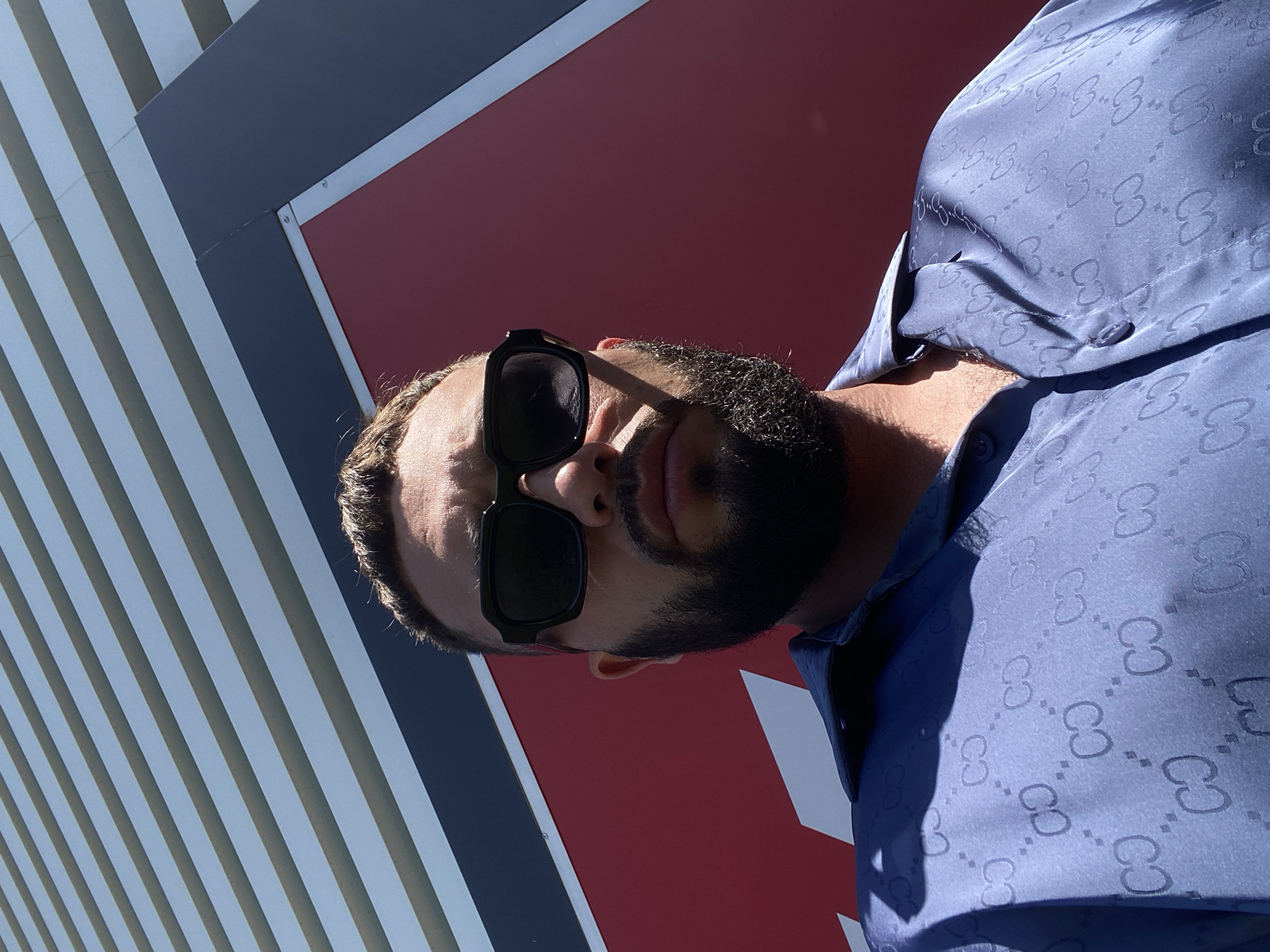 A low-angle, close-up, outdoor, eye-level selfie features a man with dark hair and a full beard wearing sunglasses and a blue button-up shirt with a repeating "GC" pattern. He is positioned in front of a large, red, angular structure with horizontal grey and dark blue stripes above it. The lighting suggests it is daytime, casting a shadow across the man's face. The image appears to have been taken in the city of Korkmaskala, Russia, given the context.