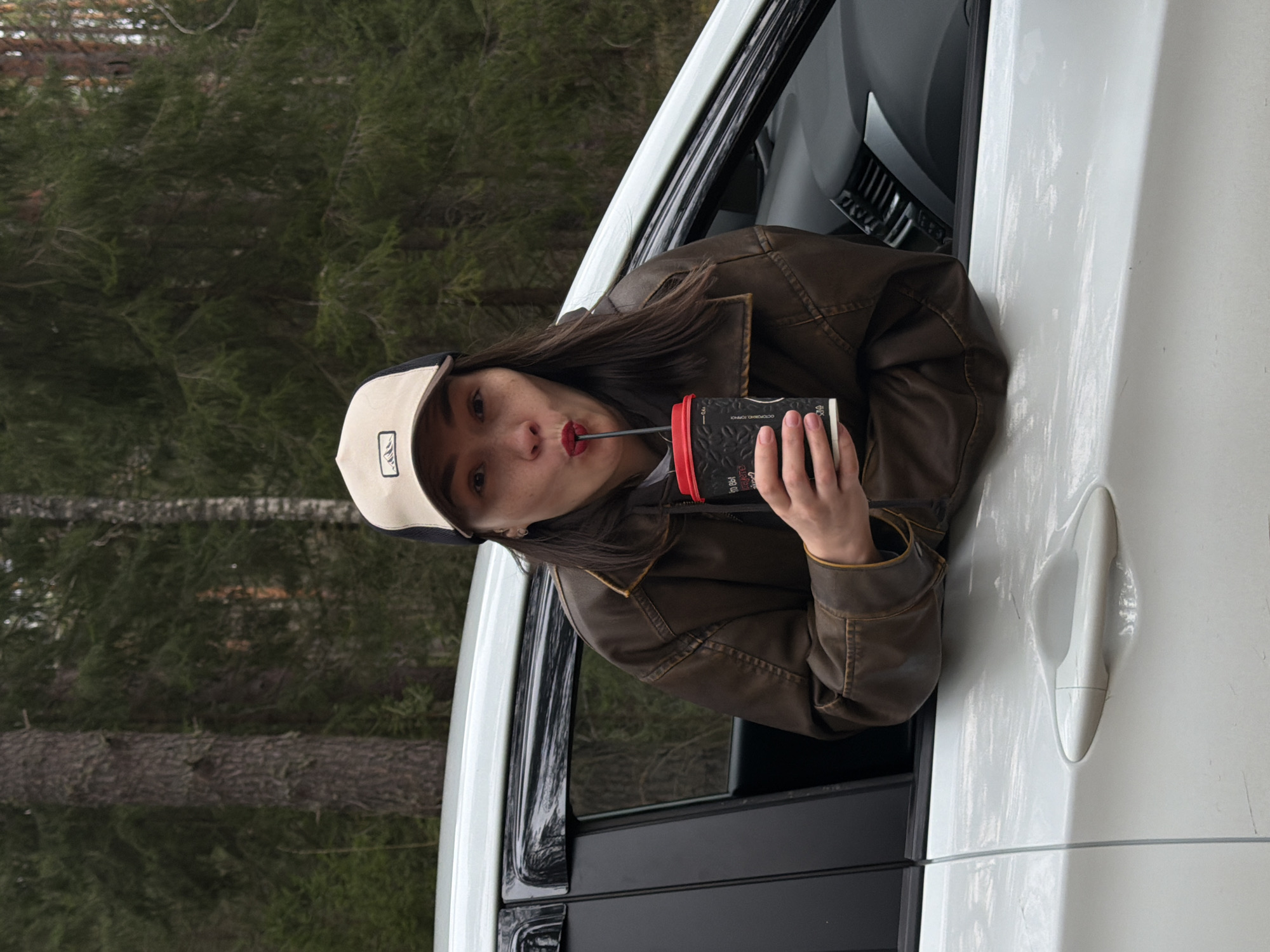 A young woman is depicted leaning out of the passenger side window of a white car, looking directly at the camera. She is drinking from a black disposable cup with a red straw, her lips pursed around it. She has long, dark hair and is wearing a brown leather-like jacket and a light-colored baseball cap with a dark brim and a small logo. Her expression is neutral, possibly concentrating on drinking.
The black cup has red accents and visible Cyrillic text, including "ОСТОРОЖНО ГОРЯЧЕ" (CAUTION HOT), as well as "100" and possibly "MUG". The white car's exterior, including a door handle, shows some light scuff marks or smudges.
The background features a dense forest composed of tall evergreen trees, suggesting a natural, wooded environment. The lighting is soft and diffused, indicating daytime, likely under an overcast sky, with no direct sunlight visible. This scene is set outdoors, potentially near Chaikovsky, Russia.