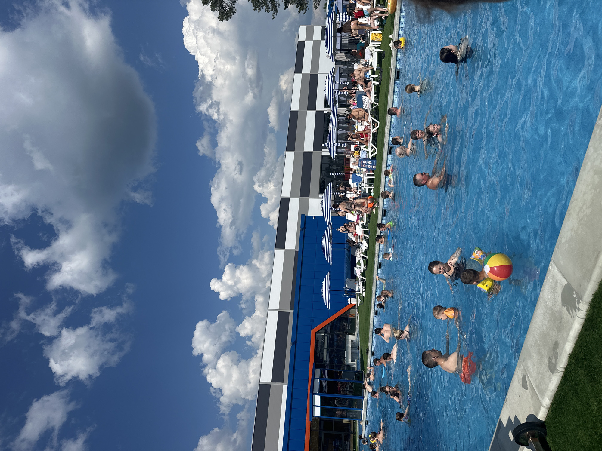 A wide, outdoor, rectangular swimming pool filled with clear blue water is the central focus of the image. The pool is populated with numerous adults and children, many of whom are swimming, wading, or standing in the water. Some individuals are interacting with each other, while others appear to be relaxing or playing. A colorful beach ball floats in the water near the foreground, held by a child wearing yellow arm floaties. Adjacent to the pool, a grassy area is visible, beyond which are lounge chairs and striped umbrellas, suggesting a resort or recreational area. In the background, a modern building with large windows and blue and grey facade panels is situated. The sky above is a vibrant blue, dotted with fluffy white clouds, indicating a sunny day. The overall scene depicts a lively summer atmosphere at a public swimming facility.