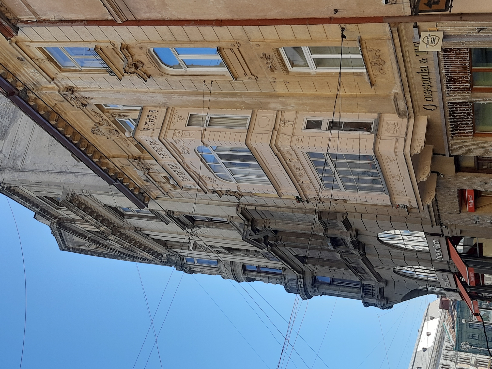 The image shows a street view of historic buildings in Lviv, Ukraine, under a clear blue sky. The architectural styles suggest an older European city. One building on the right is painted a light yellow-ochre and features ornate balconies and windows with white frames. To its left, a darker, grey stone building stands out with its classical facade, curved corner, and arched windows on the lower floors. A prominent signage for "STEFANO RICCI" is visible on this building. Further to the left, another building with a lighter stone facade and elaborate cornices is partially visible. Overhead, electrical wires crisscross the sky, a common sight in urban environments. The ground level of the buildings appears to house businesses, with awnings and shopfronts visible. The overall impression is one of a well-preserved historical urban setting.
