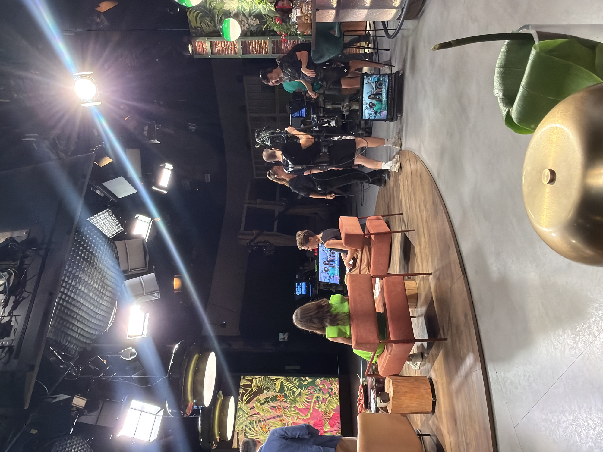 This is an indoor scene, likely a television studio, set in the city of Sant Joan Despí, Spain. The studio features a raised wooden platform with a curved edge, on which two people are seated in orange chairs. A man in a blue blazer stands to the left, facing away from the camera. In the background, a film crew is actively working. One person operates a large video camera on a pedestal, while others are positioned around them, some wearing headsets. Several monitors display video feeds. The studio is equipped with numerous overhead lights, some of which are brightly illuminated, creating lens flares. A large, colorful mural or screen is visible on the left wall. The overall atmosphere suggests a live television production is in progress.