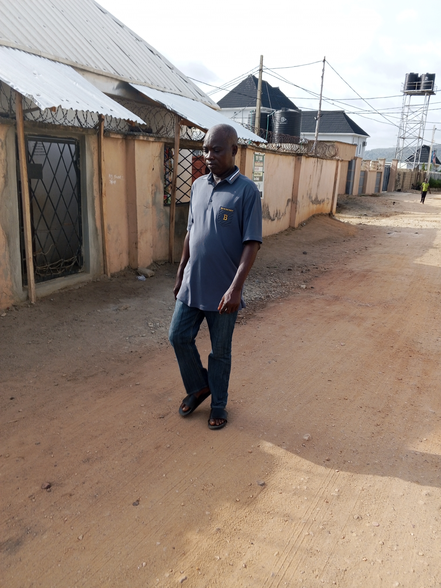 A bald, middle-aged man wearing a grey polo shirt with a Burberry logo, blue jeans, and black sandals is walking on a dirt road in Otokiti, Nigeria. To his left is a building with a corrugated metal roof and a barred window. A beige-colored wall runs parallel to the road behind the man, topped with barbed wire. Another building with a dark roof and black water tanks can be seen in the background. Further down the road, a water tower and another person in a bright shirt can be seen. The road is sandy and bare.