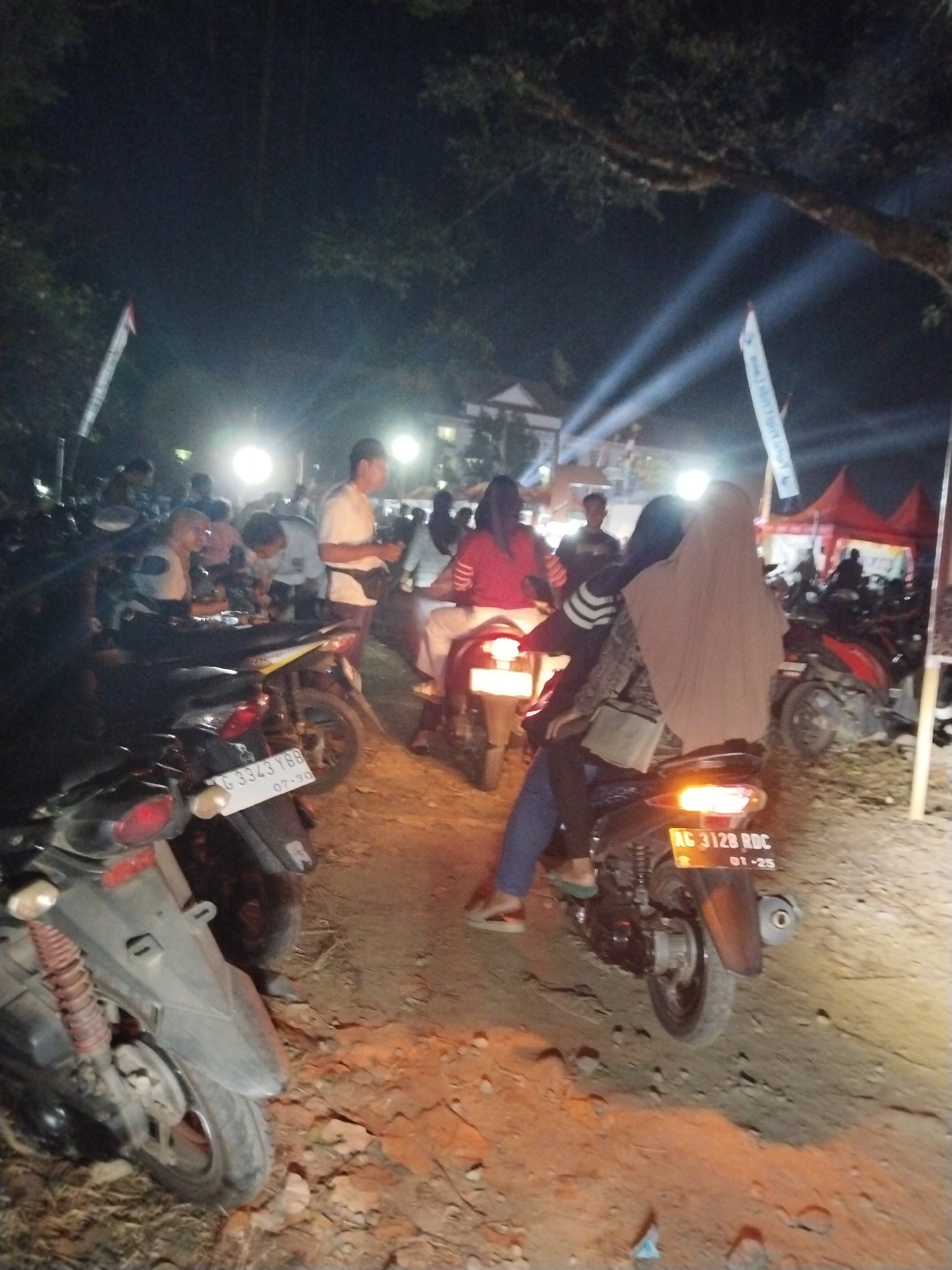 In a dimly lit outdoor scene, possibly an event in Tasikmadu, Indonesia, multiple individuals are gathered around parked motorcycles. The ground is uneven, with dirt and some debris visible. Bright lights, likely from temporary lighting setups, illuminate the area, casting long beams into the darkness. Several motorcycles are the primary focus, with some people sitting on them or standing nearby. In the foreground, a scooter is parked, its license plate "G 3343 YBB" partially visible. Further back, another motorcycle has a license plate reading "AG 3128 RDC" with "01-25" below it, suggesting an expiration date. People are dressed casually, with some wearing headscarves. One individual is seated on a motorcycle, wearing a light brown hijab and a patterned jacket, with another person behind them also wearing a hijab. Another person in the middle ground, wearing a white t-shirt, appears to be interacting with others. In the background, a red tent and more motorcycles suggest a public gathering or market. Trees with dark foliage are visible overhead.