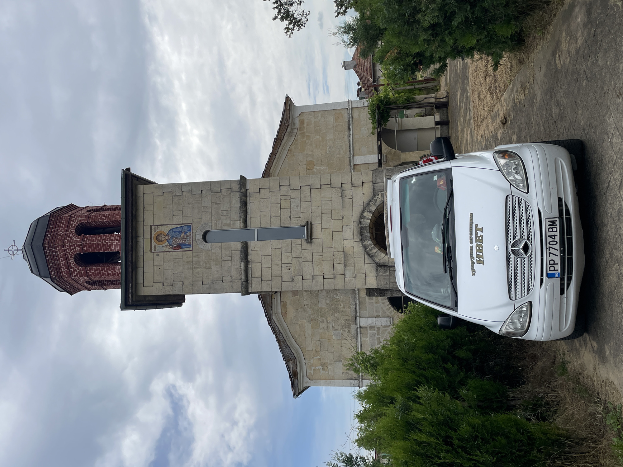 The image depicts a white Mercedes-Benz van parked near the Church of Saint Petka in Tsar Kaloyan, Bulgaria. The church is a stone building with a brick bell tower, adorned with an icon. The sky is cloudy. The vehicle has a Bulgarian license plate, "PP 7704 BM," and text on its windshield is barely visible. The van is adjacent to some greenery.