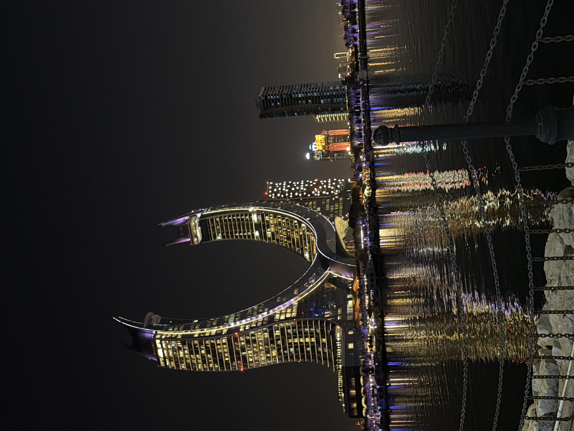 The image depicts the iconic Katara Towers in Lusail, Qatar, at night. The distinctive twin crescent-shaped skyscrapers are illuminated with purple and yellow lights, casting a warm glow against the dark sky. The buildings stand tall along the waterfront, with their reflective facades mirroring the vibrant lights. In the foreground, a calm body of water reflects the illuminated towers and the surrounding cityscape. Several boats are docked along the waterfront, adding to the picturesque scene. The image also shows a railing with chains in the lower right corner. The overall atmosphere is one of modern architectural grandeur and serene urban beauty.