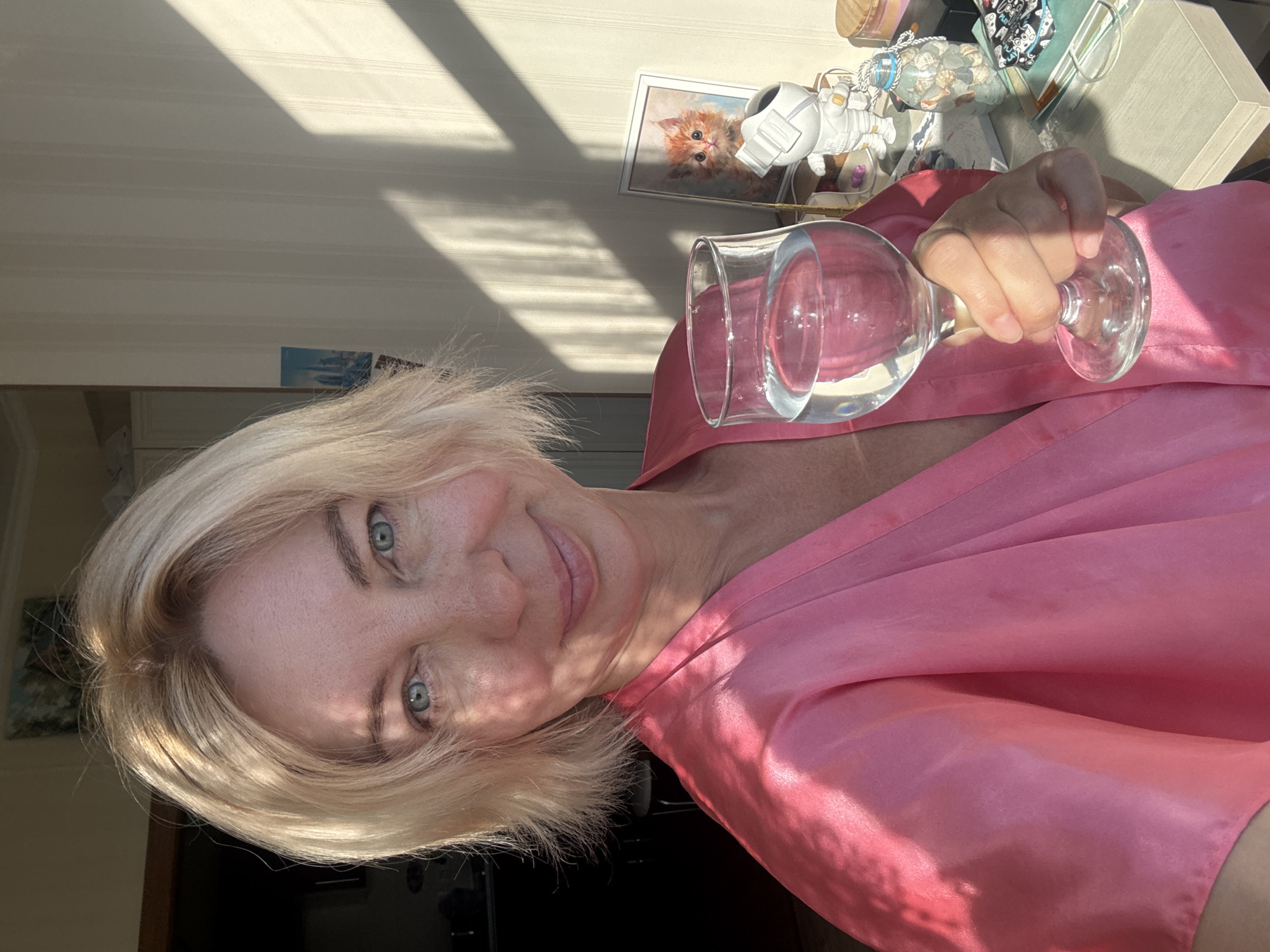 A blonde woman with blue eyes, wearing a pink silk robe, holds a glass of water. She is standing in what appears to be a room with sunlight casting shadows from blinds on the wall. On a shelf behind her, there are several decorative objects, including a framed picture of a kitten, a figurine of an astronaut, and a glass bottle filled with shells. The room is lit by natural light, creating a warm and inviting atmosphere. There is no indication of the specific location within Sochi, Russia, or any explicit interaction or action beyond the woman holding the glass.