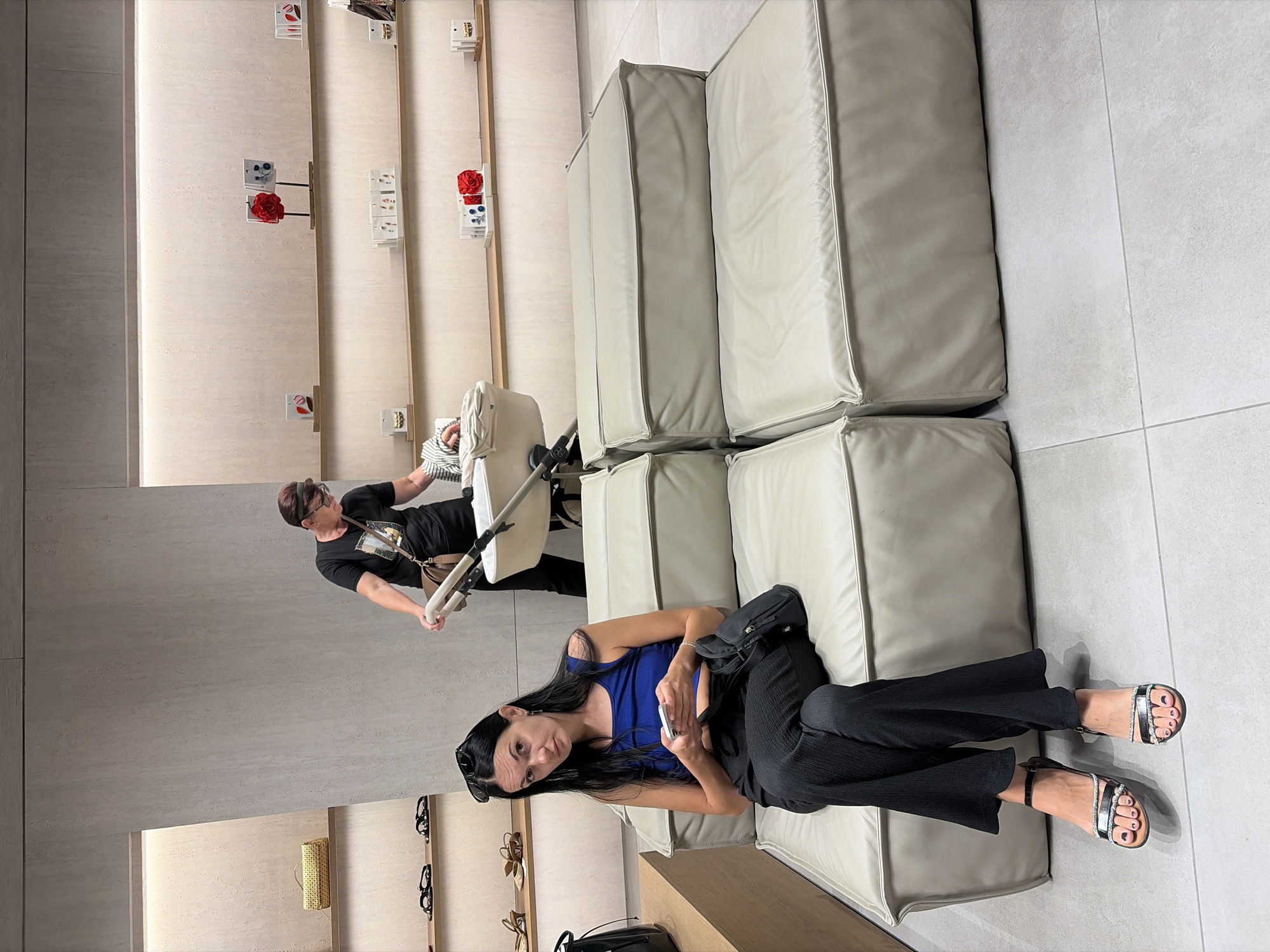 A woman with long dark hair and a blue tank top sits on a beige modular sofa in what appears to be a retail store. She is holding a smartphone and looking towards the camera with a neutral expression. Her black, wide-legged pants are pulled down to her ankles, revealing metallic sandals with jeweled straps. A black handbag rests on her lap. In the background, a woman with short dark hair and glasses pushes a baby stroller, the infant inside partially visible. The store has minimalist shelving displaying various items, including small boxes with images and red floral accents, bags, and shoes. The flooring is light-colored tile. The overall environment suggests a modern boutique or department store.