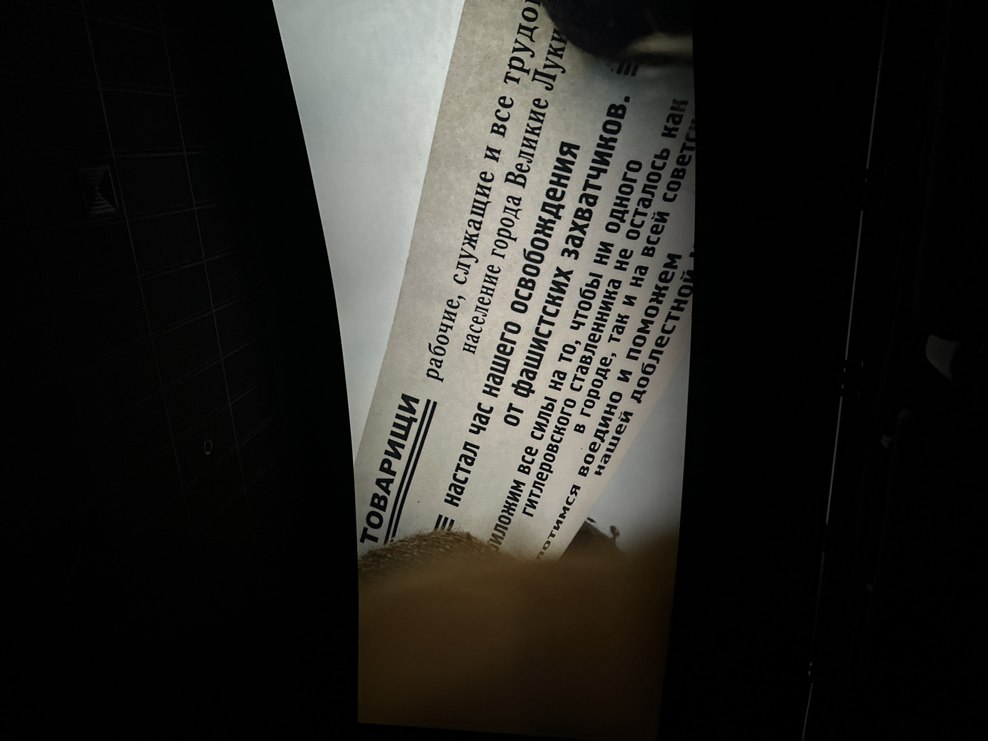 Here's a description of the image:
The image shows a piece of paper with text written in Russian. The visible text includes phrases like "Comrades, workers, employees and all laborers," "population of the city of Velikie Luki," "the time has come for our liberation," "from the fascist invaders," and "We will put all our strength to ensure that not one Hitler henchman remains in the city and on the entire Soviet land." The text appears to be part of a propaganda leaflet or announcement from the World War II era. The paper is lit diagonally, suggesting it may be lying on a surface. Some hair is visible at the bottom of the image. The picture is set against a very dark background. There's no indication that the image was taken in Krasnoyarsk, Russia.