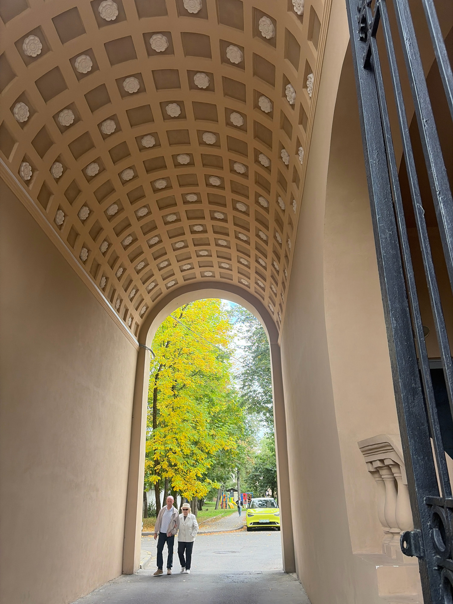 This is an eye-level, medium shot captured outdoors, looking through an arched passageway. The passageway ceiling is decorated with a grid of recessed squares, each containing a white floral embellishment. The walls of the passageway are a light tan color. To the right, a portion of a black metal gate is visible.
Beyond the arch, a street is visible with a backdrop of trees displaying vibrant yellow and green autumn foliage. Two individuals, an older couple, are walking arm-in-arm away from the camera, on the paved pathway. The man is wearing a light-colored jacket and dark pants, while the woman is in a white jacket, dark pants, and a light-colored hat. Further down the street, a bright yellow compact car is parked, and in the distance, a playground structure is partially visible.
The scene is located in Minsk, Belarus, specifically at the entrance to the courtyard of the National Art Museum of the Republic of Belarus.