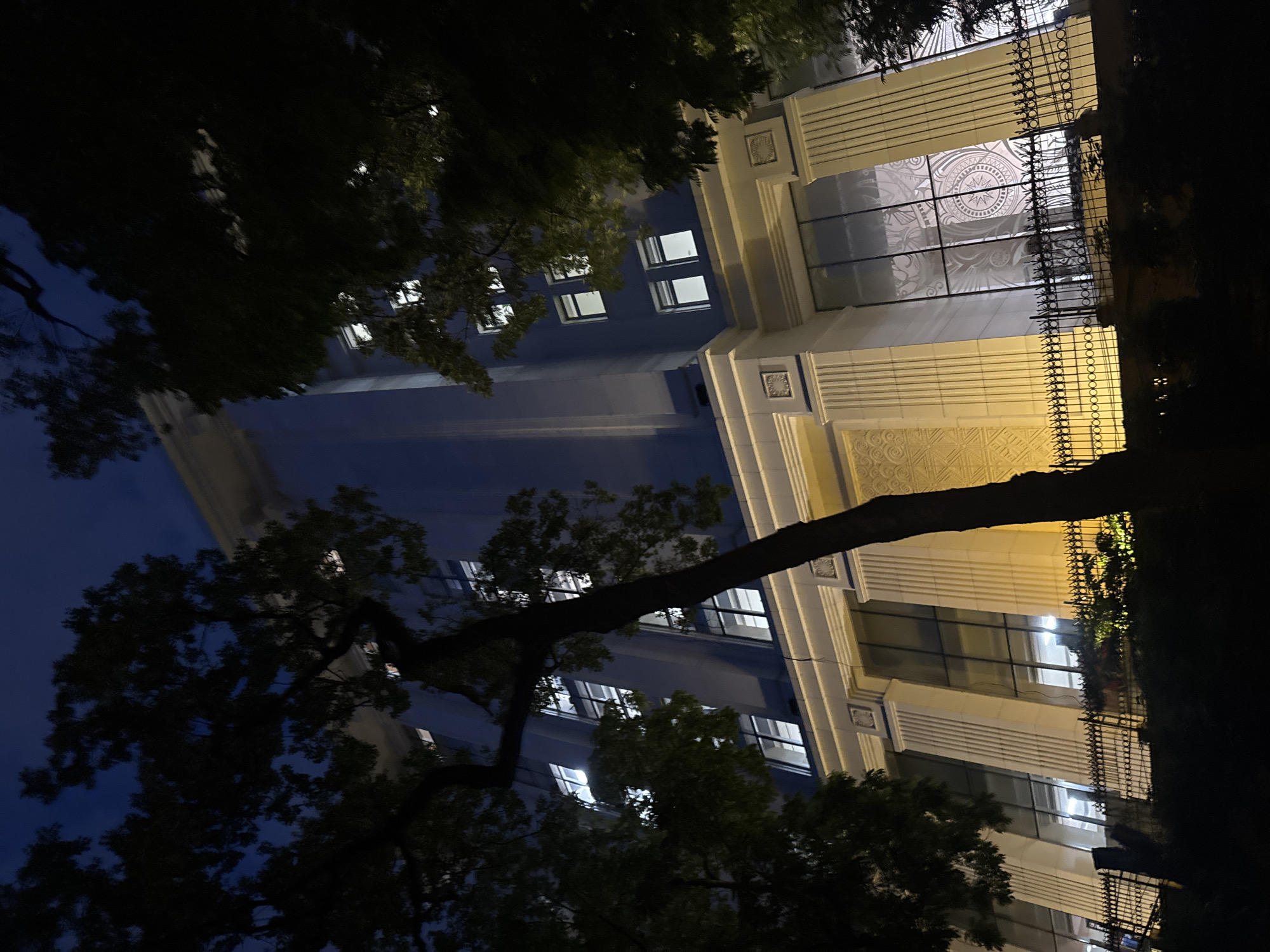 The image captures a nocturnal scene in Hanoi, Vietnam, focusing on a building with illuminated windows and decorative architectural elements. The building's facade features classical columns and patterned sections, suggesting a historical or grand structure. A black metal fence with intricate scrollwork borders the building at the lower right, with some greenery visible behind it, possibly from a balcony or garden.
Silhouetted trees with dark foliage dominate the foreground and upper left portion of the image, their branches reaching across the frame. The contrast between the dark trees and the lit windows of the building creates a dramatic effect. The time of day is clearly night, indicated by the darkness of the sky and the artificial lighting. The weather appears calm, with no visible signs of rain or strong winds.
The image is composed with a low-angle perspective, looking upwards towards the building and trees. The building's windows are uniformly lit with a warm, white light, suggesting interior spaces are occupied or well-lit. The reflections on the building's glass surfaces hint at the presence of other lights or elements in the surrounding environment, though these are not clearly discernible. There is no visible text in the image. The overall mood is serene and slightly mysterious, with the interplay of light and shadow adding depth to the scene.