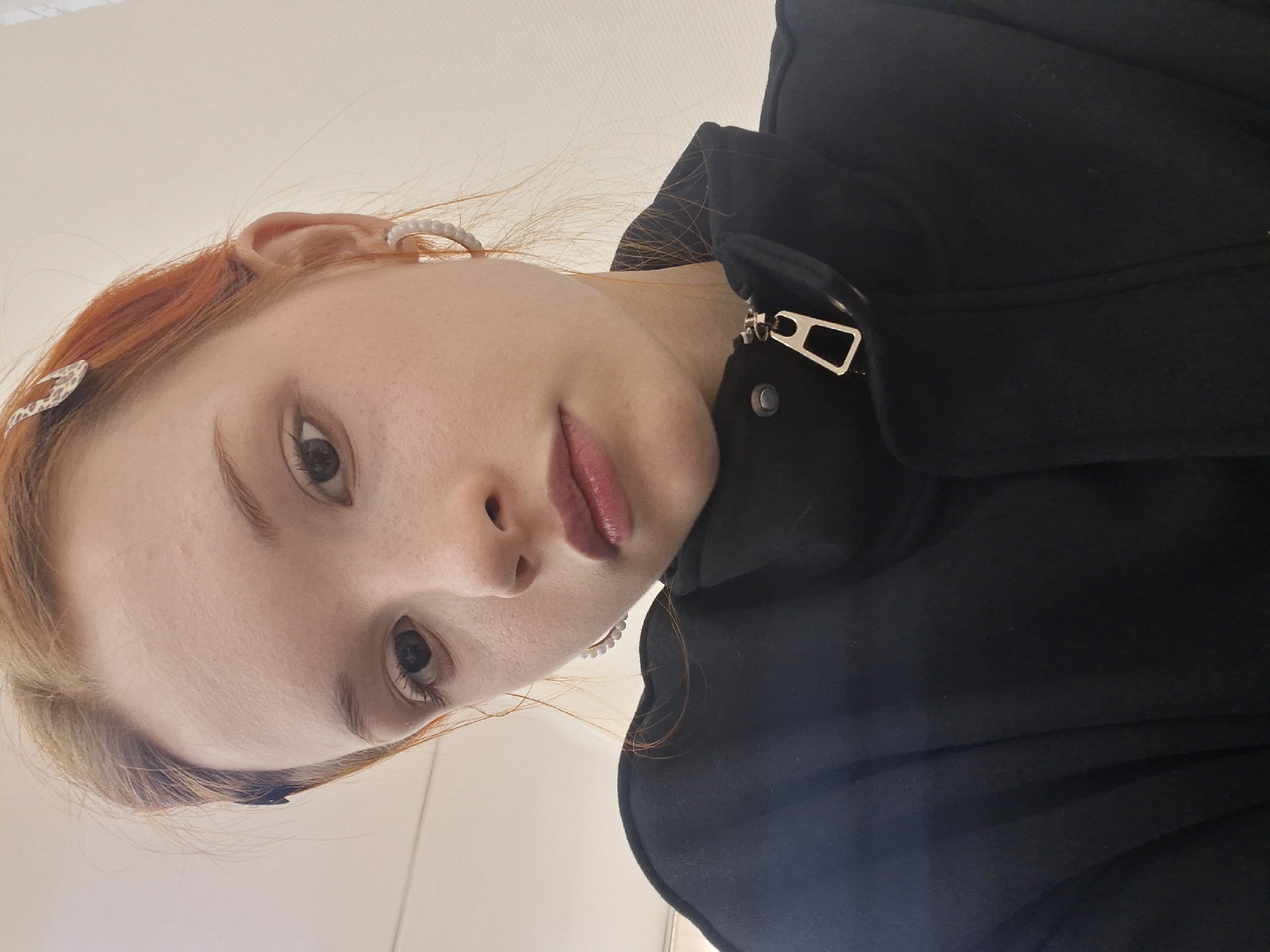 A low-angle portrait captures a young woman with reddish-blonde hair and fair skin, looking directly at the camera. Her hair appears to be parted, with lighter blonde tones framing her face. She has light-colored eyes, and her lips are painted with a soft rosy-berry hue. Subtle freckles are visible on her nose and cheeks. Her hair is adorned with two clips: one featuring a leopard print or similar pattern on her left side, and another with small white beads or pearls positioned on her right side. A beaded or pearl earring, matching the hair clip, is visible in her right ear. She is wearing a black jacket or coat, with a distinct silver-toned zipper pull visible near the collar. The background is a plain, light-colored wall or ceiling, indicating an indoor setting. Her expression is neutral and direct, suggesting she is posing for the photograph. No text is visible in the image. The location context provided is Chaikovsky, Russia.