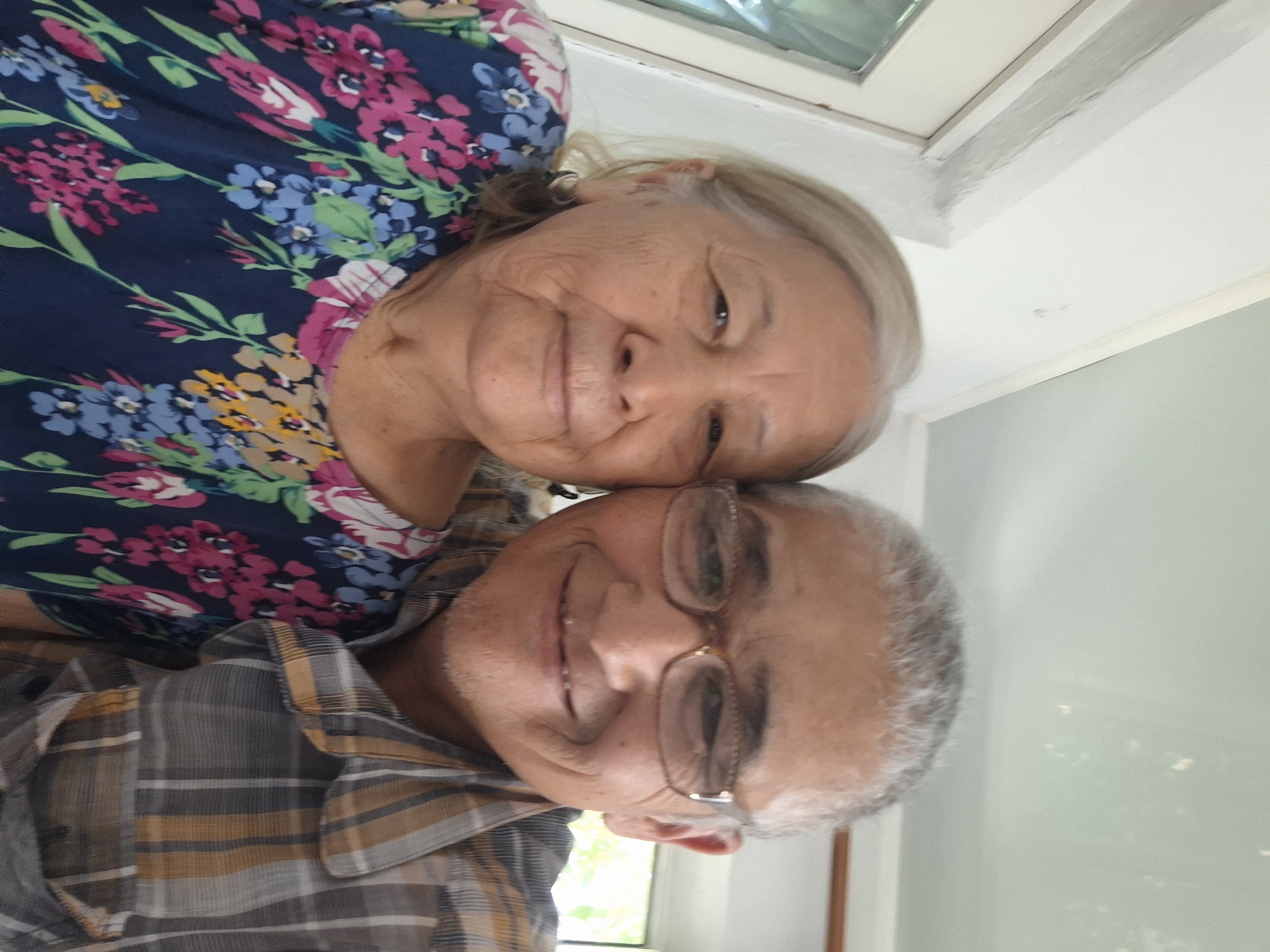 A selfie of an elderly woman and an elderly man, likely a couple, is captured in a close-up outdoor shot. The woman, positioned on the left, has fair skin, grey hair, and is wearing a dark blue top with a vibrant floral pattern featuring pink, blue, and yellow flowers. She is looking directly at the camera with a gentle smile. The man, on the right, has fair skin, grey hair, and is wearing glasses with light brown frames. He is also smiling at the camera and is dressed in a plaid shirt with gray, yellow, and white stripes. They are standing close together, with the man's arm likely around the woman, though only their upper torsos and faces are visible. The background suggests an outdoor setting with natural light, possibly a balcony or patio area, with a hint of greenery visible through a window or open space. The lighting is soft and diffused. The image does not provide specific visual cues to definitively pinpoint the location in the city of Almaty, Kazakhstan.