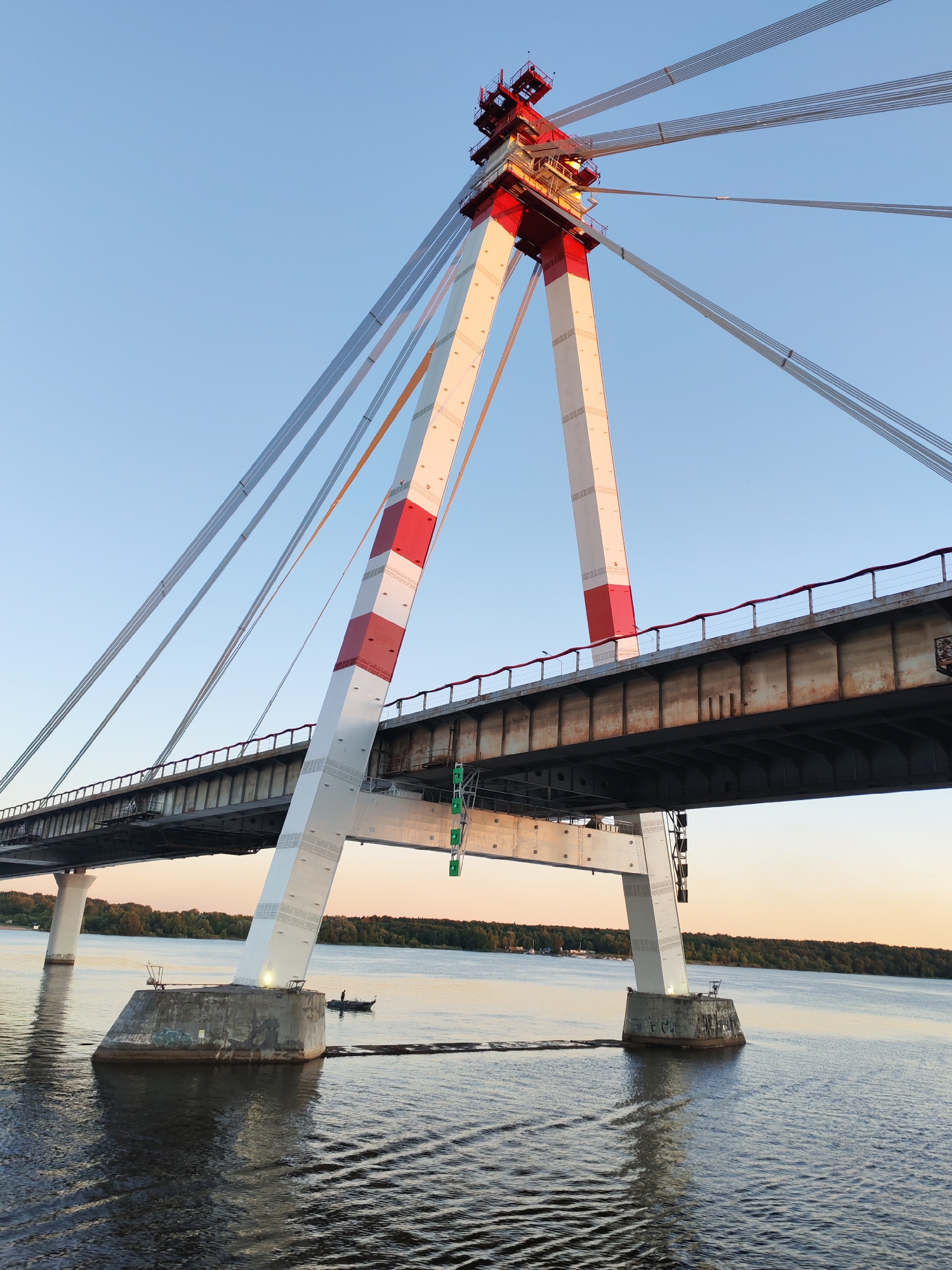 The image shows a cable-stayed bridge over a body of water, likely a river, with a clear blue sky. The bridge has two large white pylons with red markings, supporting a roadway. The water has ripples, and a small boat with a person in it is visible near the shore. In the distance, a treelined bank is visible. The bridge appears to be the Yuryevetsky Bridge in Cherepovets, Russia, which spans the Rybinsk Reservoir on the Sheksna River. The sun is low in the sky, suggesting it is either early morning or late evening.