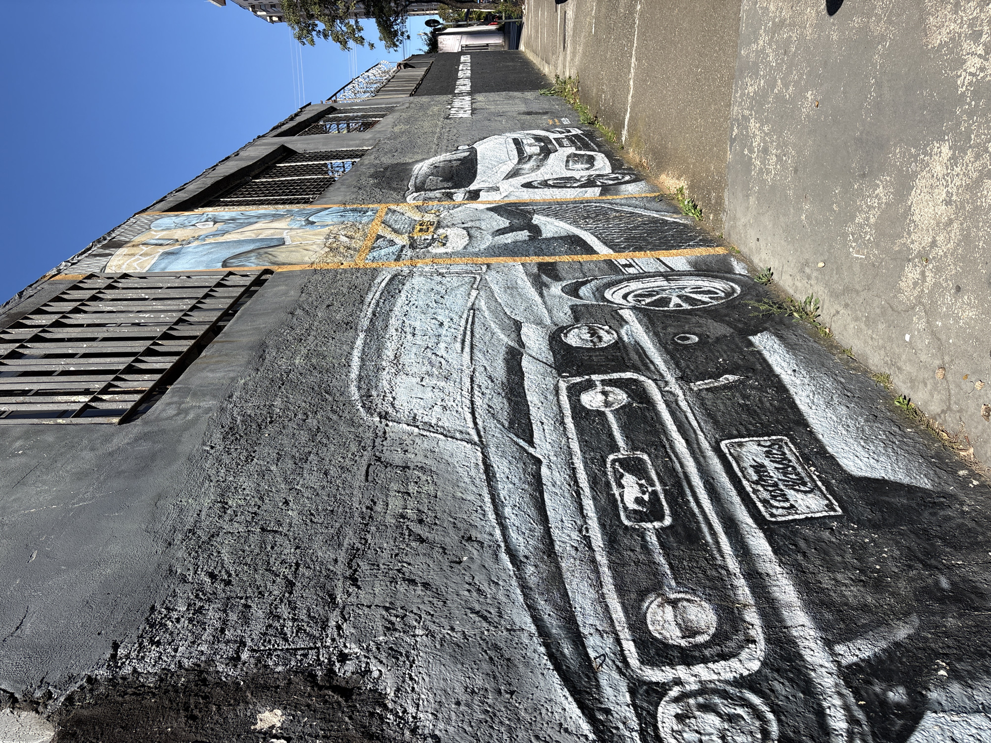 The image shows a mural on a textured, grey concrete wall, depicting two cars. The car in the foreground is a classic black and white painting of a Ford Mustang with a "Custom Classics" license plate and a horse emblem on the grille. To its right, a white modern car is also depicted. The mural extends down the length of the wall, which is bordered by a sidewalk and a patch of grass. Above the mural, barred windows are visible on the wall, and a clear blue sky dominates the upper portion of the image. The wall and sidewalk show signs of wear and tear, with some cracks and debris. The location appears to be a street in São Paulo, Brazil, as indicated by the text on a sign visible further down the street, which reads "MECÂNICA".