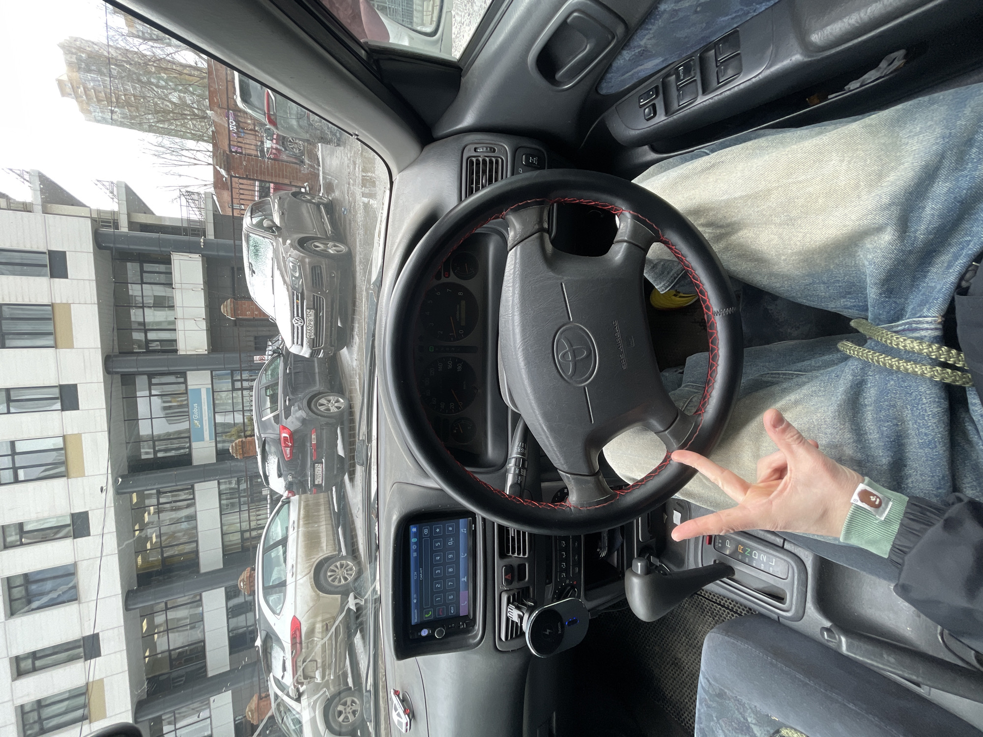 The image shows the interior of a car, specifically the driver's side. The steering wheel features a Toyota logo and is wrapped with red stitching. The dashboard includes a screen displaying a phone dial pad, and a device is plugged into the cigarette lighter. The gear shift is visible. The person in the driver's seat is wearing jeans and a jacket with a patch on the wrist and is holding up their hand in a gesture. Through the windshield, parked cars are visible, as well as a building with "Globus" sign. Based on the visible license plates, the building sign, and the architecture of the building outside the window, the location is likely within the city of Krasnoyarsk, Russia.