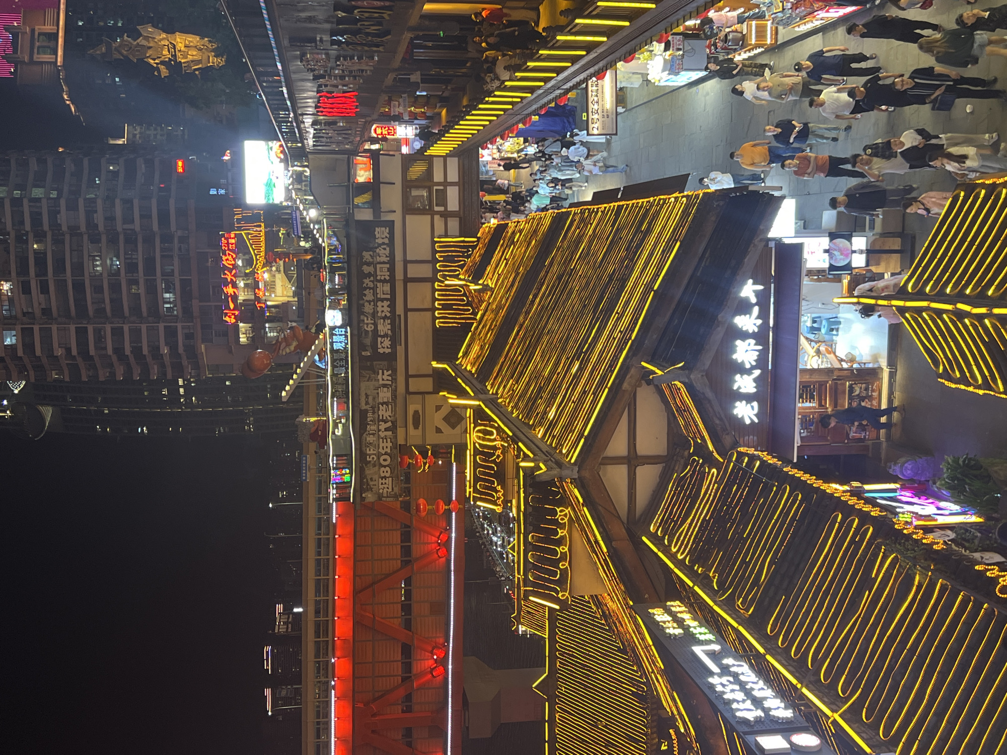 The image captures a vibrant night scene in Yuzhong District, China. The focal point is a traditional Chinese building with multiple layers of rooftops, extensively decorated with bright yellow neon lights that trace the rooflines. A red bridge structure stands beside the building, with similar linear lighting emphasizing its design.
Pedestrians populate a walkway that stretches beyond the building, with shops or stalls lining the path, creating a bustling atmosphere. In the background, modern high-rise buildings dominate the skyline, lit up with various neon signs. The signs are adorned with Chinese characters and contribute to the lively urban environment.
Visible text indicates the presence of restaurants and tourist attractions, like "Lui Yishou Big Pot" and "Hongyadong Panorama Lookout", hinting at the location's popularity. The overall scene depicts a blend of traditional architecture and modern urban life, illuminated by an array of vibrant lights.