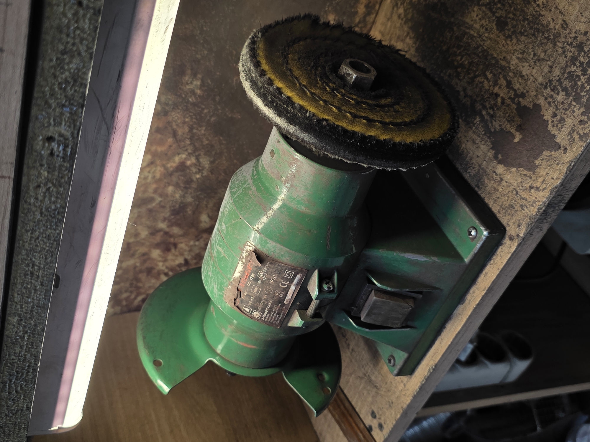The image shows a green bench grinder with a polishing wheel attached, resting on a wooden workbench. The bench grinder is positioned on the right side of the frame, with its polishing wheel facing towards the viewer. The machine appears to be old and well-used, with some rust and wear visible on its surface. The wooden workbench is rough and weathered, showing signs of use and age. Above the bench grinder, a fluorescent light fixture illuminates the scene. The background is a textured wall, possibly made of wood or concrete, with a mottled brown and gray appearance. No people are visible in the image. The scene suggests a workshop or a place where tools are used for maintenance or craftsmanship. The image does not contain any elements that would indicate its specific location in Kazan, Russia.