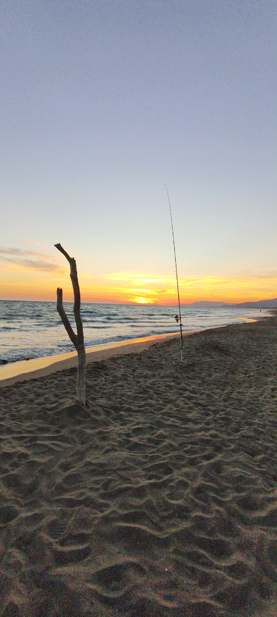 The image depicts a tranquil beach scene at sunset. The sand in the foreground is dark and textured, with ripples created by the wind or water. A piece of driftwood, with two distinct branches, stands upright in the sand. To the right, a fishing rod with a reel is planted in the sand, its tip angled towards the sea. The sea itself is visible, with gentle waves lapping the shore. The horizon is marked by a line of hills or mountains in the distance, silhouetted against a vibrant sunset sky. The sky transitions from a pale blue at the top to shades of orange and yellow near the horizon, where the sun is setting. There is a faint, narrow strip of people visible walking along the shoreline in the distance.
While the image is a beautiful depiction of a sunset on a beach, it does not provide any specific visual cues to pinpoint its location within the city of Grosseto, Italy. Beaches in the Maremma region of Tuscany, where Grosseto is located, often feature similar characteristics, but without additional context or identifying landmarks, a precise location cannot be determined.