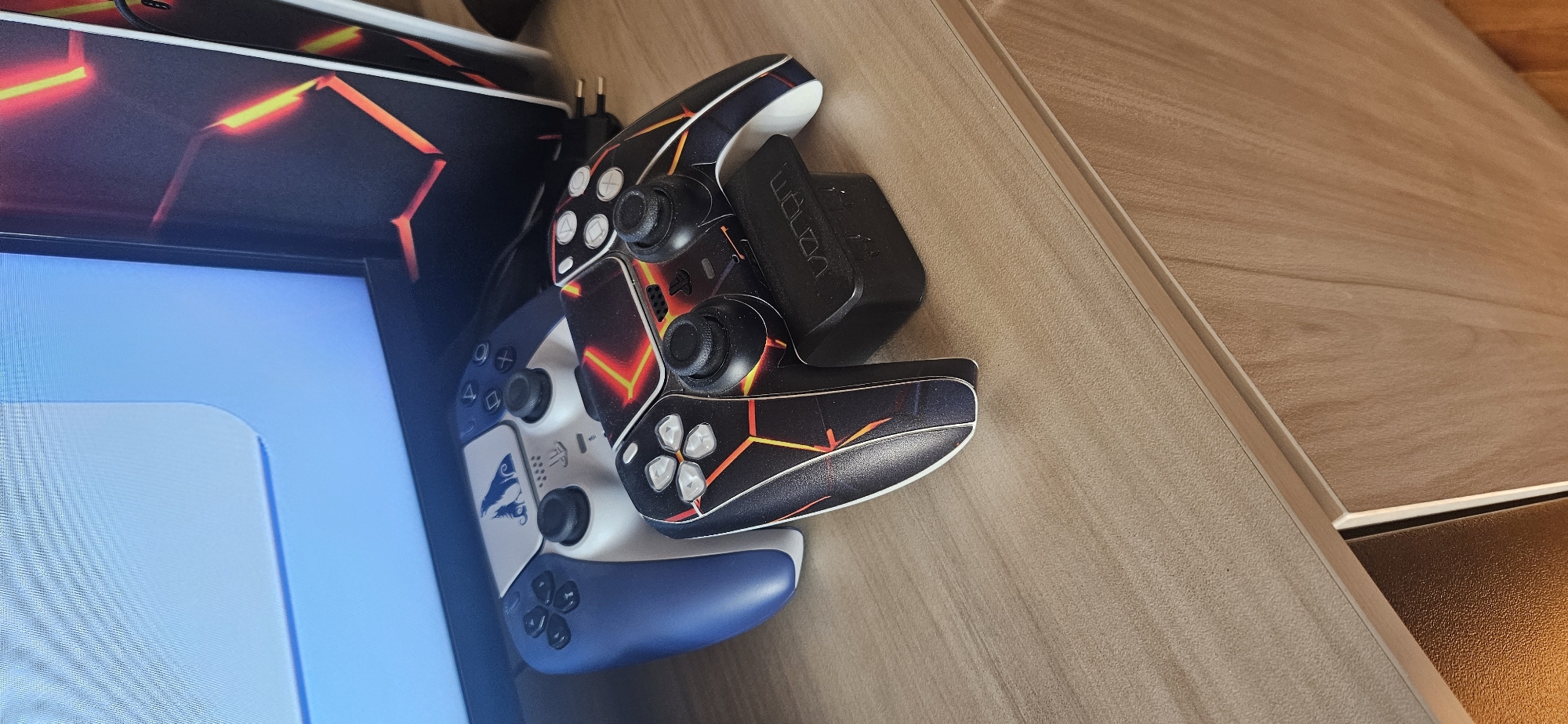 On a light brown wood-grain table sits a PlayStation 5 console partially visible on the left, and two PlayStation 5 DualSense controllers. One controller has a dark skin with red-orange lightning-like patterns, and is connected to a Venom charging dock. The second controller is blue and white with a stylized wolf design. The console's side panel is also skinned with a similar dark skin with red-orange patterns. The scene is well-lit and the items appear to be indoors. The specific location in Sofia, Bulgaria, is not discernible from the image.