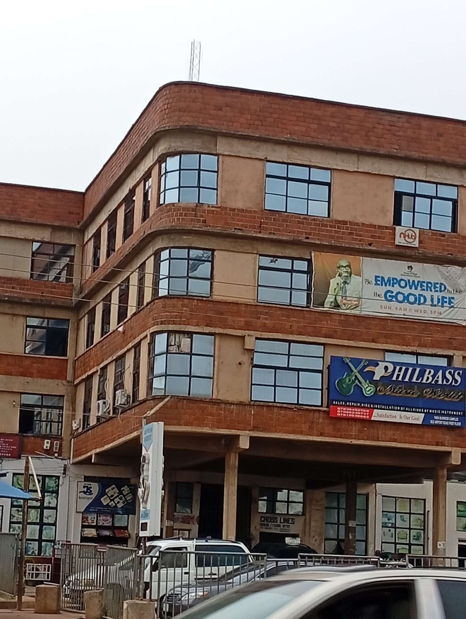 The image shows a multi-story building with a reddish-brown brick facade and beige stucco walls. The building features numerous rectangular windows with dark frames, many of which are gridded. The architectural style appears somewhat dated, with rounded corners on some sections of the building.
Several signs are visible on the building. One large banner depicts a man in glasses and text that reads "Be EMPOWERED to the GOOD LIFE SUN, 8AM // WED, 5PM". Another prominent sign, dark blue with green accents, displays an image of guitars and reads "PHILBASS Music World". Below this, smaller text lists services and a slogan: "Satisfaction Is Our Goal". A smaller sign with a logo that appears to be "nHUB" is also present.
At street level, there are storefronts with display windows. One sign reads "CROSS LINES". Another sign is a vertical white pylon with text on it, some of which is obscured. Various vehicles are visible in the foreground, including a white van and parts of other cars, suggesting it is a public street or parking area.
The environment appears to be urban. The sky is overcast, indicating a cloudy day. The building's design and the presence of commercial signs suggest it is a business or mixed-use development. The question also mentions the city of Jos, Nigeria, which provides context for the location of this building.