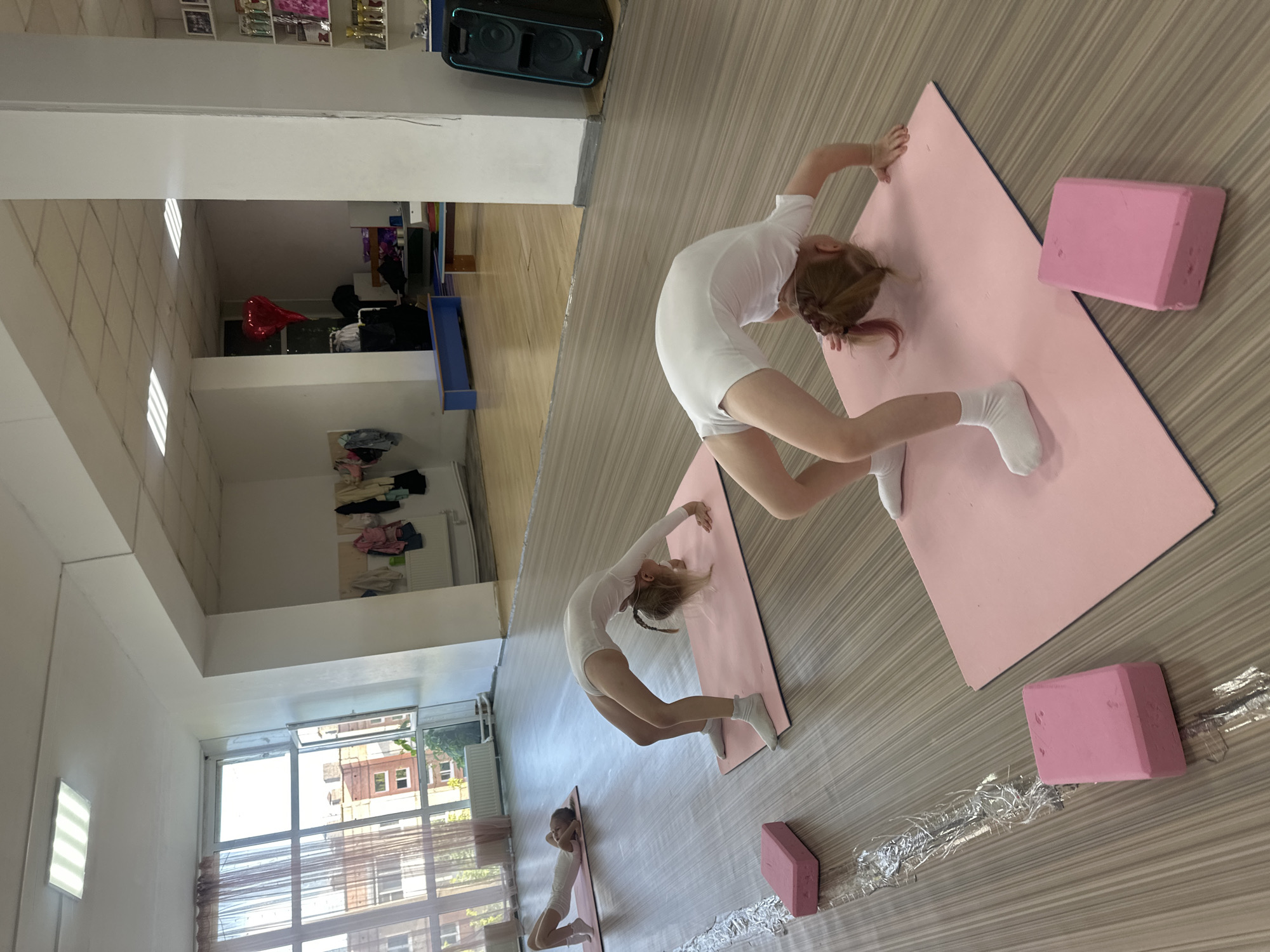 In a gymnasium located in Votkinsk, Russia, three young girls in white leotards are practicing gymnastics on pink yoga mats. The girl in the foreground is performing a backbend, with her hands and feet on the mat. Another girl behind her is also in a backbend position, while a third girl further in the background is lying on her back. Pink yoga blocks are placed on the floor next to the mats. The room has wooden flooring and large windows with sheer curtains, allowing natural light to enter. In the background, there are shelves with various items and a speaker system. A red heart-shaped balloon is visible hanging from the ceiling. The overall scene depicts a children's gymnastics class.