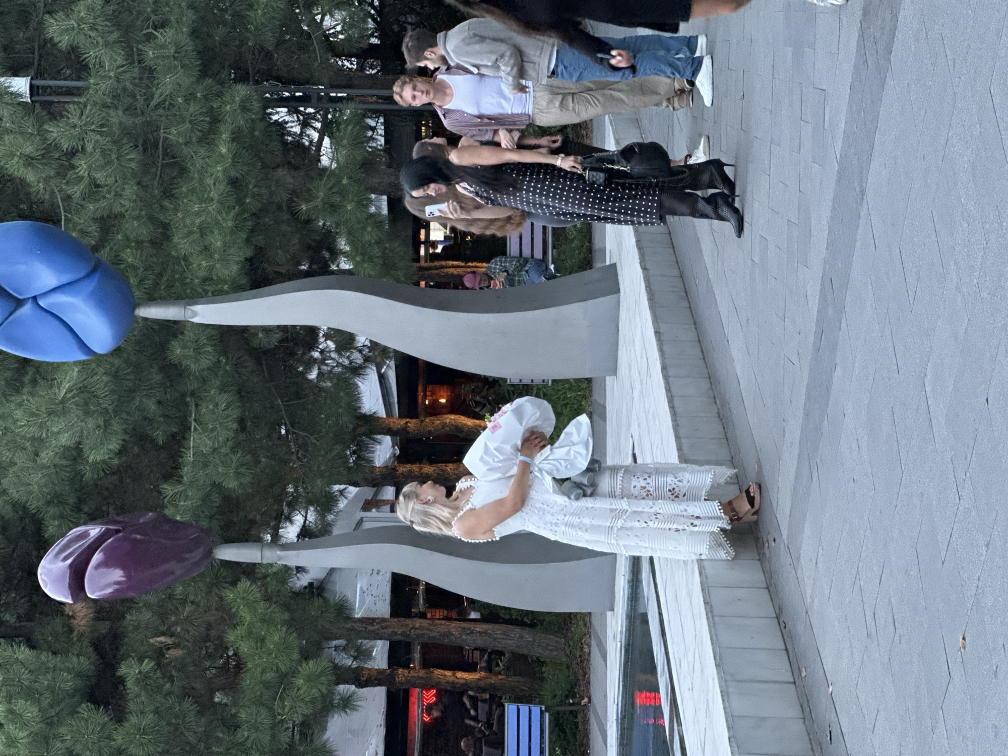 The image depicts an outdoor public space, possibly a plaza or park, with large, abstract sculptures resembling stylized flowers. In the foreground, a woman in a white lace dress stands by a water feature, holding a bouquet wrapped in white paper. To her right, a group of people are gathered. One woman in a black polka-dot dress is taking a photo with her phone. Behind her, a man in a white tank top and striped shirt is talking with another man in a light-colored jacket and jeans. The background shows trees, buildings with illuminated signs, and benches. The overall setting suggests a contemporary urban environment. The location is not Odesa, Ukraine.