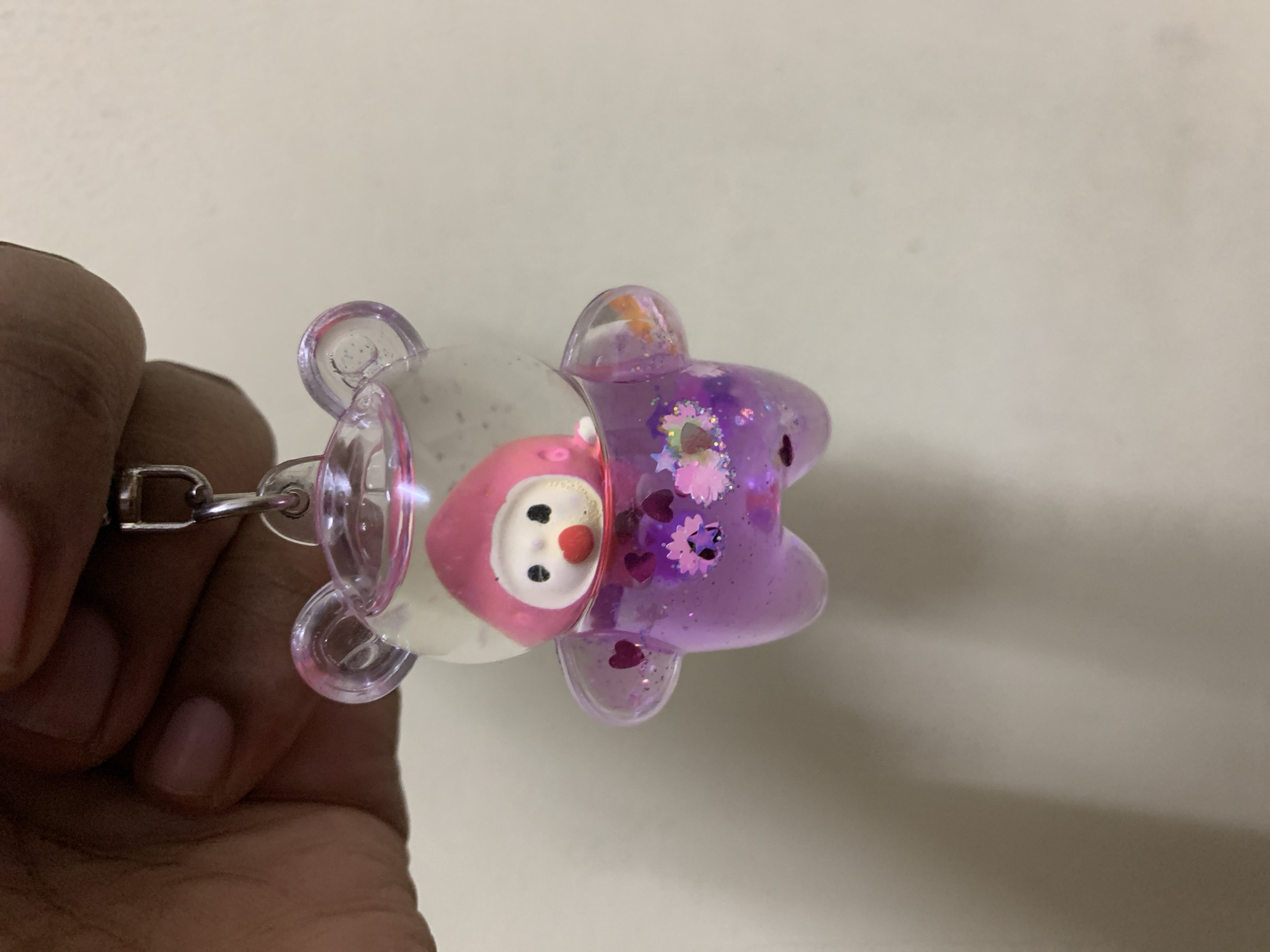 A hand holds a translucent bear-shaped keychain with a pink and white figure inside the front section and purple liquid with glitter and heart-shaped confetti in the rear section. The background is a solid light beige. The location is not discernible from the image.