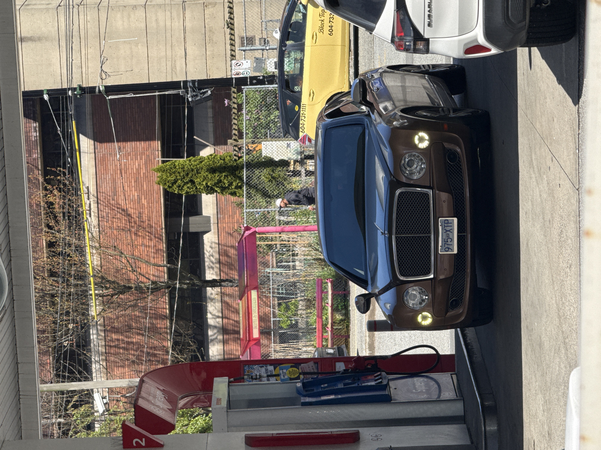 The image depicts a gas station in Vancouver, Canada, featuring an Esso pump with a brown Bentley parked in front of it. To the right of the Bentley is a white Subaru, and further back, a yellow taxi. The background shows a building with a mix of brick and concrete walls, along with various utility lines and a tree. A person is visible between the Bentley and the building. The scene is captured on a sunny day. The license plate of the brown Bentley is visible showing the number "975-XTP".