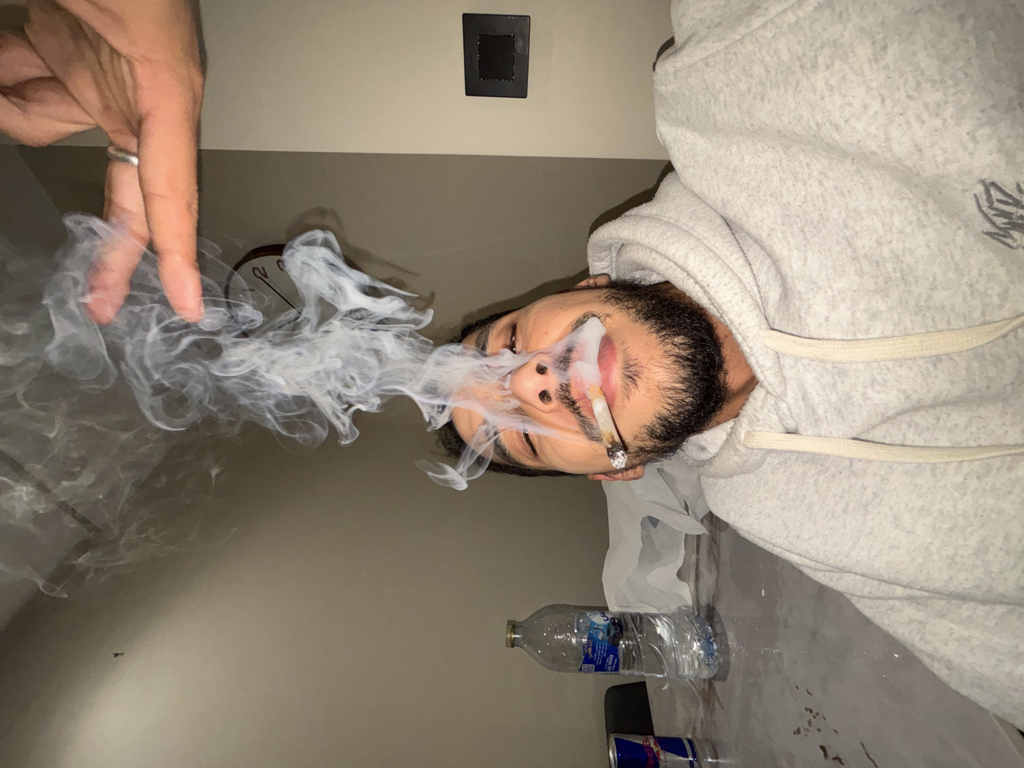 The scene is identified as taking place indoors in the city of Tangier, Morocco.
A male individual is depicted from the mid-chest up, centrally positioned and looking slightly upwards. He has a dark beard and mustache and is wearing a light grey hooded sweatshirt with a light-colored drawstring and a dark graphic on the front. A lit cigarette is held between his lips, from which a substantial plume of white smoke rises upwards and to the left. His eyes appear partially closed.
To the left of the subject, on a light-colored, speckled tabletop, rests a clear plastic water bottle with a blue and white label, next to a partially visible red and blue Red Bull energy drink can.
In the upper right portion of the image, a light-skinned human hand is visible, positioned above the main subject's head, with fingers slightly splayed. A silver-colored ring is worn on the ring finger of this hand. The hand appears to be interacting with or positioned near the rising smoke.
The background consists of plain light beige walls. A partial view of a wall-mounted clock with a dark rim and a light face is visible behind the smoke in the upper left. A black rectangular electrical outlet or switch plate is present on the right wall. The tabletop exhibits some dark marks or stains.
