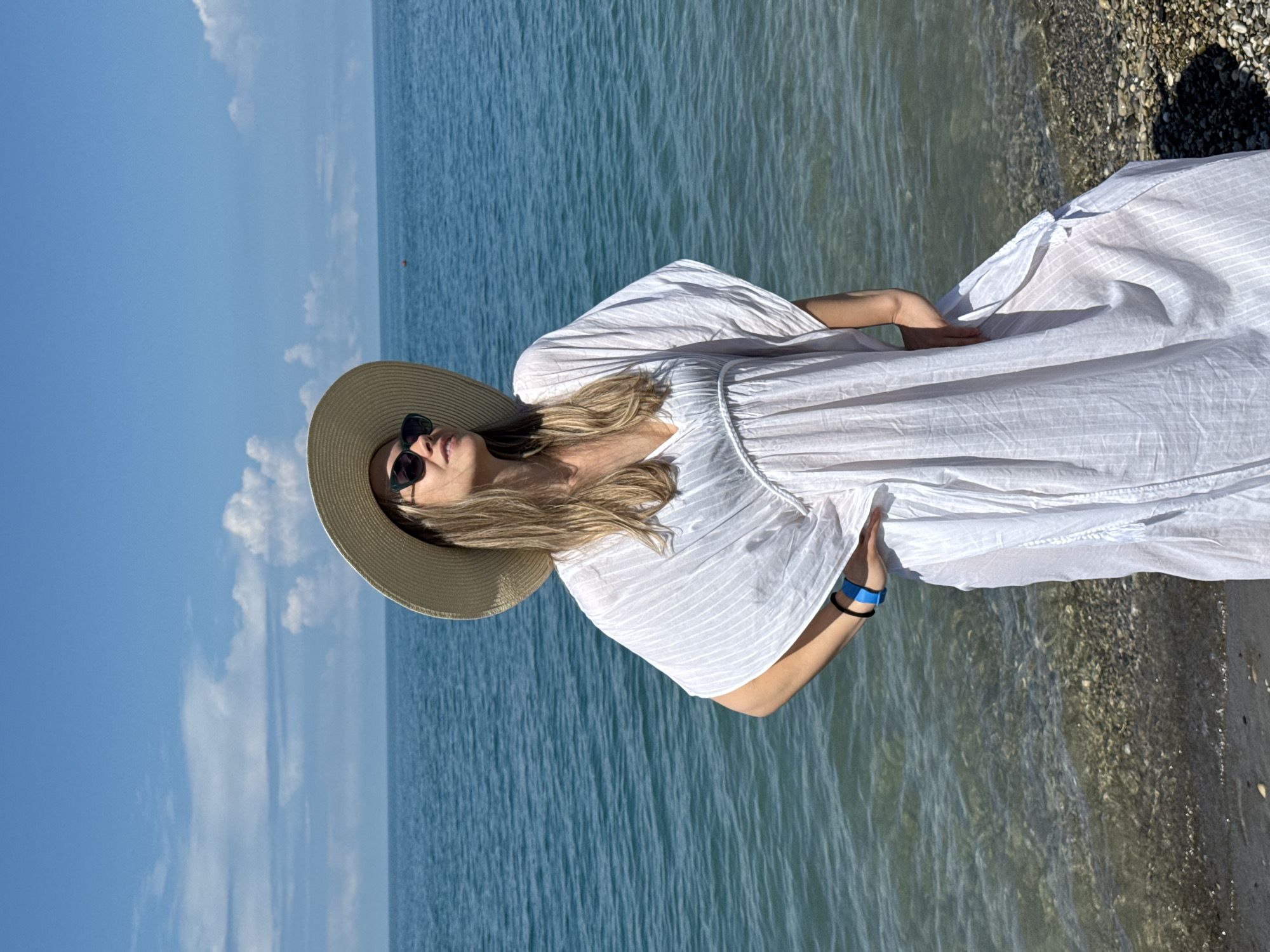 A woman stands on a pebbly beach, her body angled to the right, facing the water under a clear blue sky with scattered clouds. She wears a wide-brimmed straw hat and dark sunglasses, her blonde hair cascading over her shoulders. Her dress is a loose, white, flowing garment with vertical stripes and a V-neckline, cinched at the waist. She has one hand on her hip, and a blue bracelet is visible on her left wrist. The sea, a deep blue, stretches to the horizon, with small waves lapping at the shore. The location is identified as Nebug, Russia.