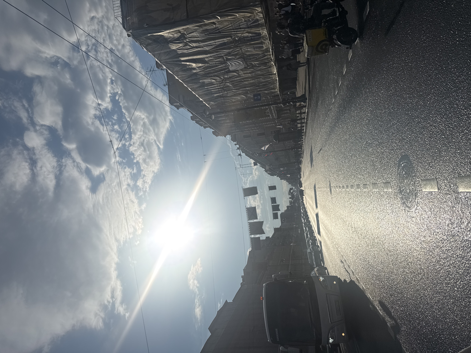 A wide, wet asphalt street in Saint Petersburg, Russia, is depicted under a bright, glaring sun and partly cloudy sky. The sun's rays create a lens flare and cast long shadows. Buildings line both sides of the street, many with elaborate facades, and one on the right is covered in protective sheeting. Traffic, including cars and a bus, moves down the street. Flags or banners are strung across the road overhead. On the right side of the street, a person on a motorized scooter with a yellow cargo box is visible. The overall scene suggests a bustling urban environment on a sunny day.