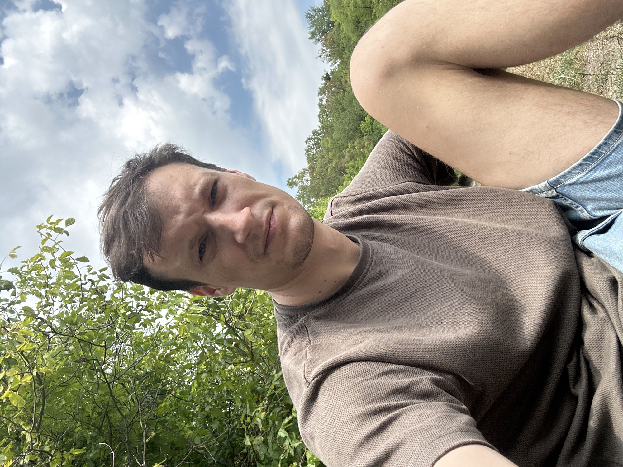 A medium shot captures a young man sitting outdoors, with his body angled toward the camera and his head tilted down. He has fair skin, short brown hair, and is wearing a brown textured t-shirt and denim shorts. His gaze is directed slightly away from the viewer, with a neutral expression. The background features lush green trees and a partially cloudy blue sky. The man's right leg is bent and resting in front of him, with his left arm positioned out of frame to the left. The setting appears to be a park or natural area, possibly within the city of Makiv, Ukraine.
