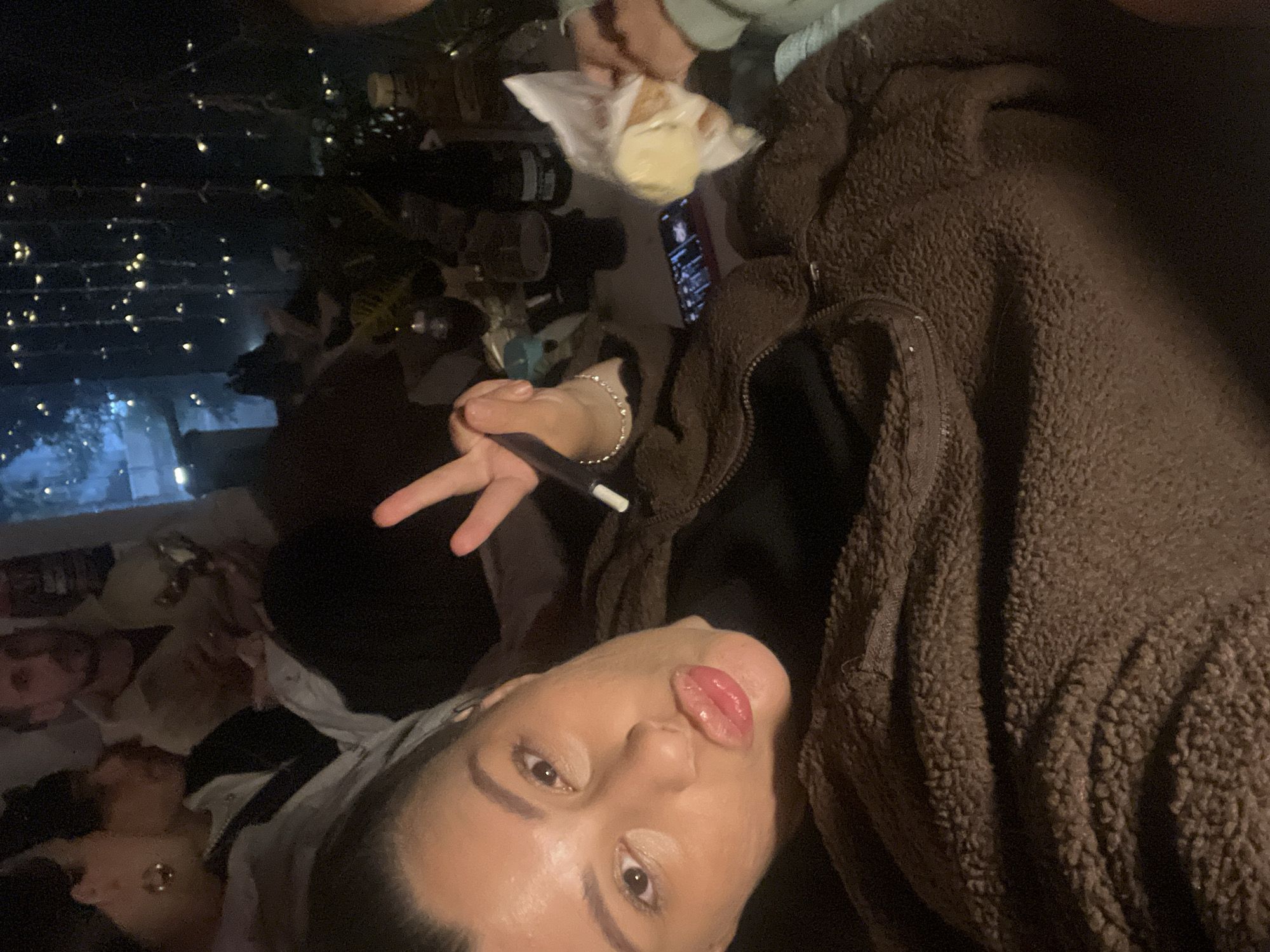 The image appears to be a selfie taken indoors, likely at night based on the lighting conditions. The foreground features a person posing with their tongue out and making a peace sign with their fingers. They are wearing a brown, textured jacket. In the background, there are several other individuals, some seemingly lying down. On a surface near the foreground, there's a cell phone, some food in a plastic bag, and bottles of liquid. String lights are visible in the background, suggesting a festive or decorative setting. The exact location within Kiryat Bialik, Israel, cannot be determined without additional contextual information.