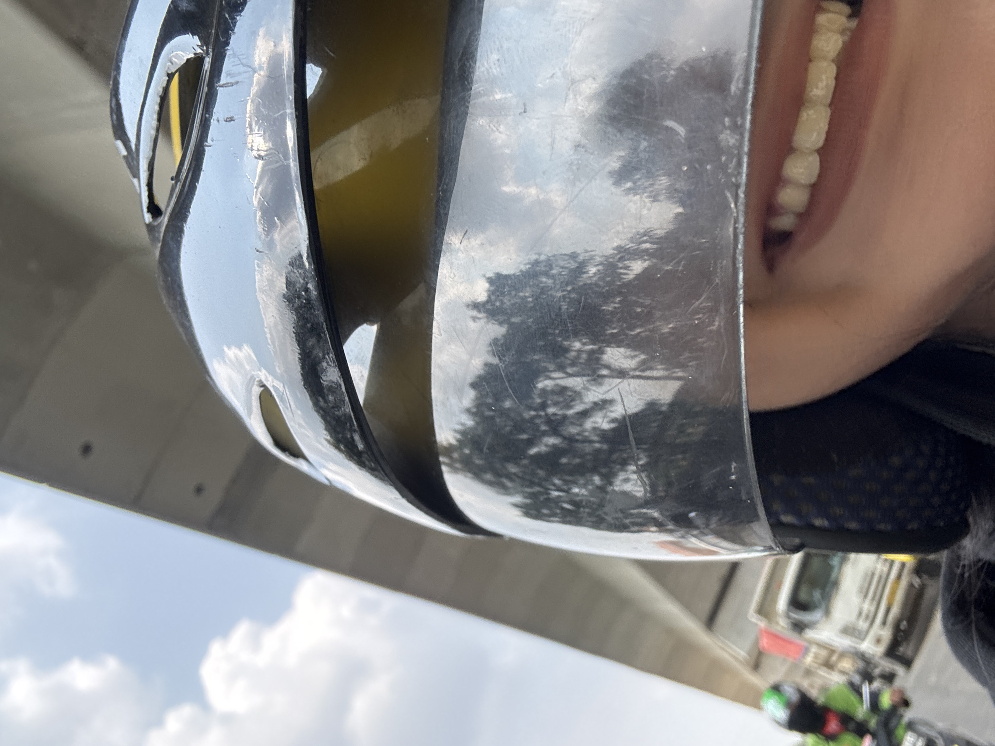 The image, taken from a low angle, shows a person wearing a bicycle helmet. The helmet is silver and has a reflective surface that mirrors the sky, including clouds and what appears to be the silhouette of trees or buildings. Part of the person's face is visible, revealing a set of teeth. The background includes a gray structure, possibly a bridge or overpass, and a glimpse of the sky with clouds. Additionally, a blurry object resembling a vehicle or camera mount attached to the bike can be seen. Based on the instructions, it is specified that the location is North Jakarta, Indonesia, though this detail is not immediately obvious from the visual information presented in the image.