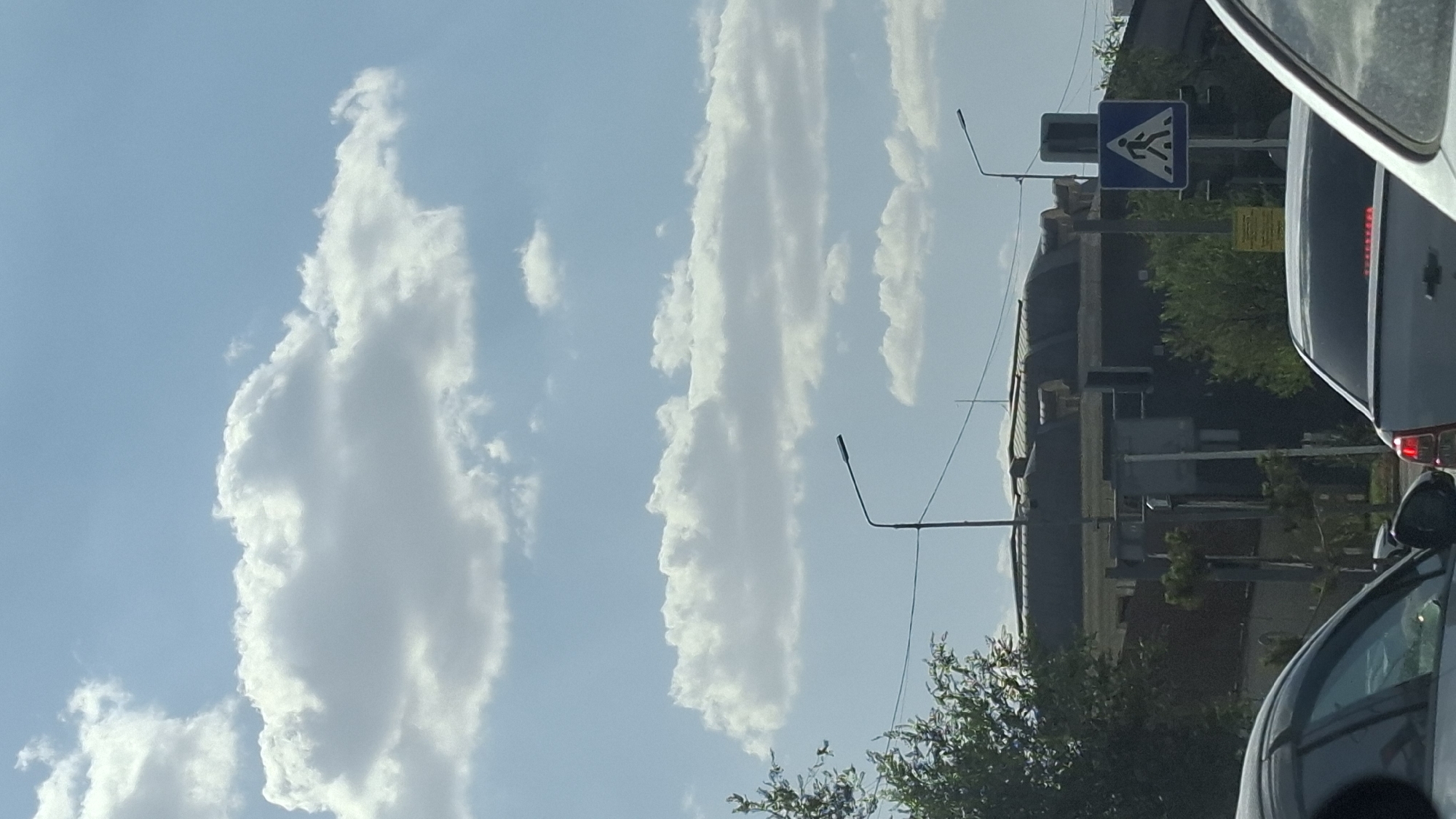 The image captures a view of a city street in Shymkent, Kazakhstan, on a clear day with a bright blue sky and scattered white clouds. In the foreground, parts of two cars are visible, suggesting they are part of a traffic queue. A pedestrian crossing sign, depicting a person walking across a road, is prominently displayed on the right side of the frame. Behind the cars and sign, buildings with dark roofs are visible, as well as trees and streetlights. The overall scene depicts a typical urban environment during daylight hours.