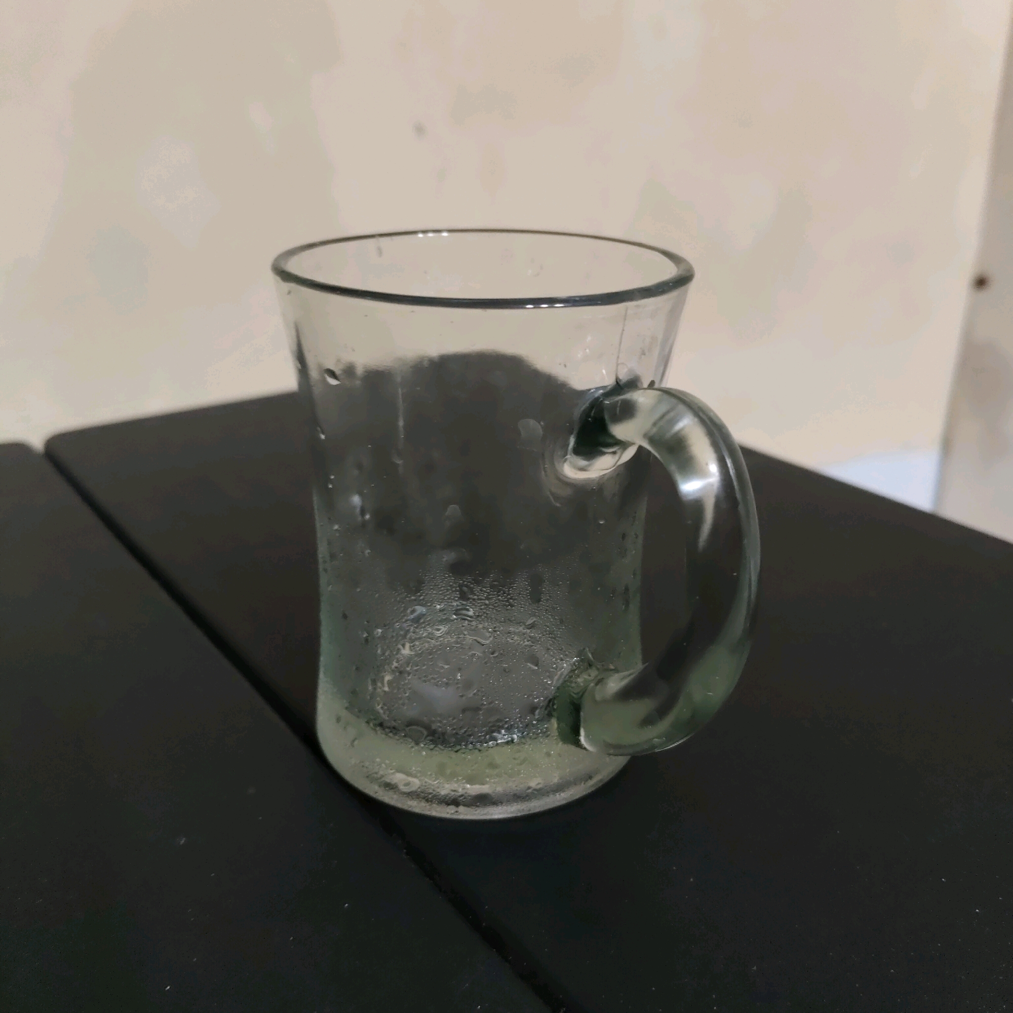 A clear glass mug with condensation on its exterior sits on a dark, textured surface. The mug has a rounded handle. The background is a plain, light-colored wall with some faint shadows. The scene is static and appears to be indoors. There are no people or actions depicted. This image was taken in Sunggal, Indonesia.