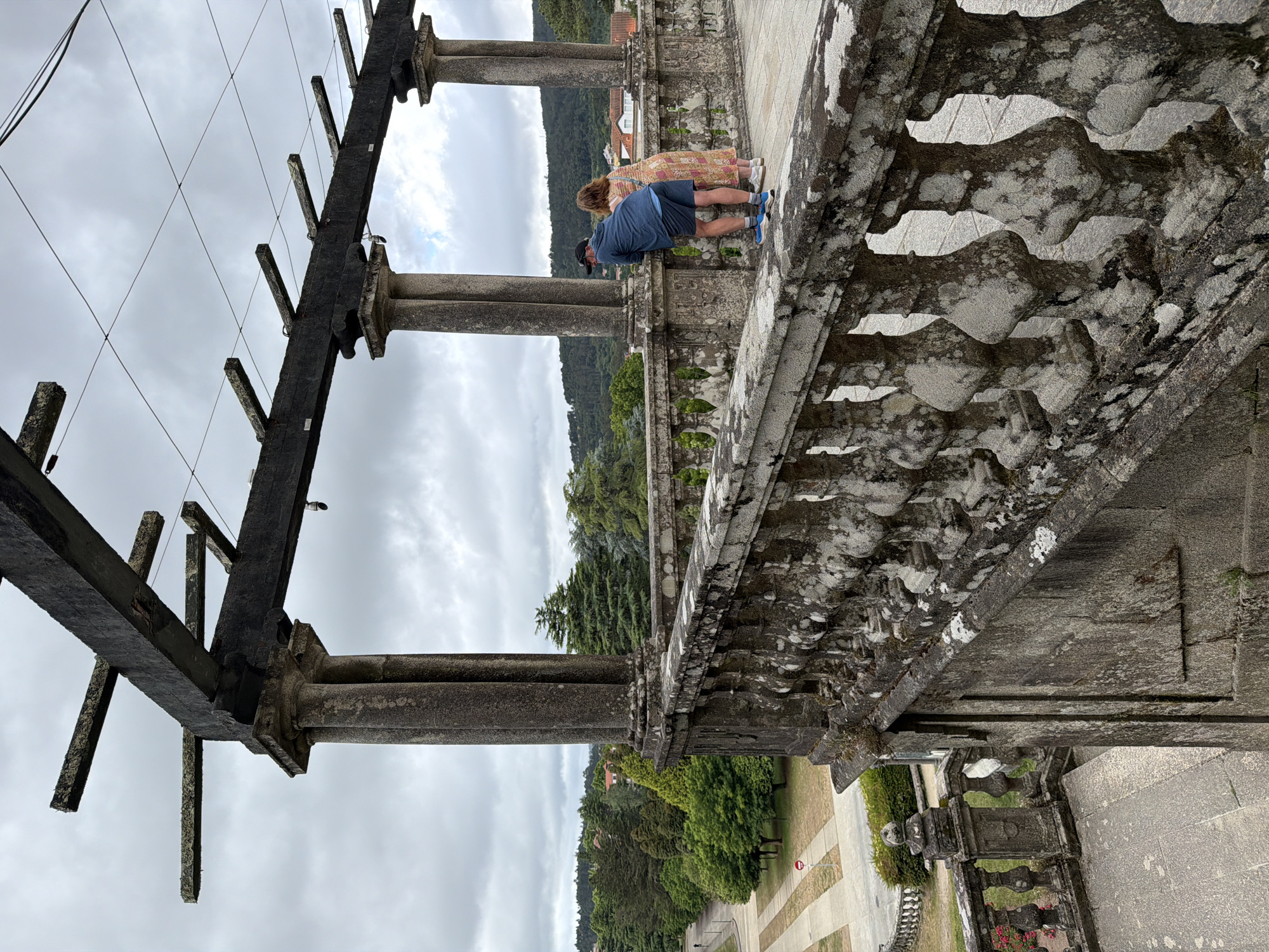 The image shows a couple from the back, a man and a woman, leaning on a stone balustrade. The man is wearing a dark blue hoodie, blue shorts, and blue sneakers. The woman is wearing a long, multi-colored floral dress and white sneakers. They appear to be looking out at the view.
The balustrade is made of stone and has a weathered, mossy appearance. Behind the couple, there is a large stone pergola structure with columns and wooden beams supporting a wire grid. The background features a lush green landscape with trees, rolling hills, and some buildings in the distance. A paved pathway and a street are visible in the lower left portion of the image, with a small traffic sign. The sky is overcast with clouds.
This is the Panteón de San Martiño de Mondoñedo, located in Santiago de Compostela, Spain. It is part of the San Martiño de Mondoñedo Cathedral complex, a historic religious site.