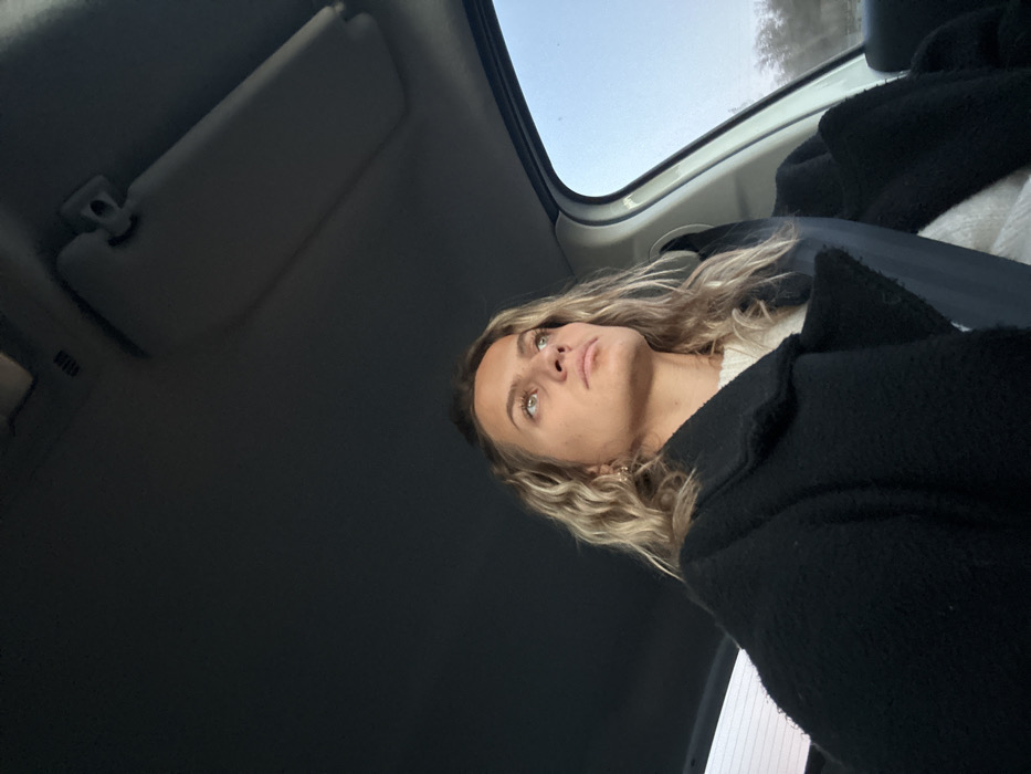 A person with blonde, wavy hair and light-colored eyes looks upward in a car. The person is wearing a white shirt under a dark, textured coat. The car's interior is visible, including a sun visor and a window showing a glimpse of the outside sky. The angle is from below, giving an upward perspective. The setting suggests travel or being a passenger in a vehicle.