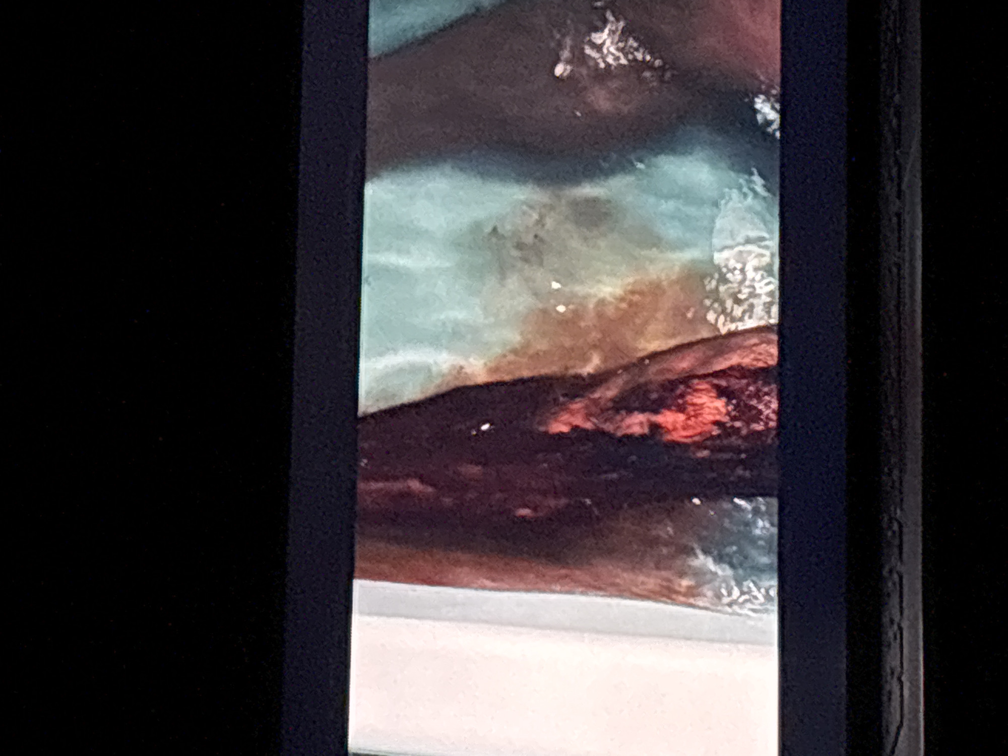 The image displays a still frame presented on an electronic screen, characterized by distinct black borders on the top and bottom. The central content depicts a close-up of what appears to be a section of a human or animal body, exhibiting a large, deep, and irregular open wound.
The wound itself features exposed tissue that is bright red and textured, indicative of muscle or subcutaneous layers. Surrounding the wound, the tissue is discolored with large areas of dark reddish-brown, consistent with dried blood or extensive bruising. To the immediate left of this central subject, a vertical light grey or white surface is visible, possibly an interior wall or architectural element. To the right and upper areas of the frame, there are mottled patches of teal and brownish discoloration, with several scattered lighter specks. The entire scene is presented without any visual indicators or contextual clues pertaining to the city of Budva, Montenegro.