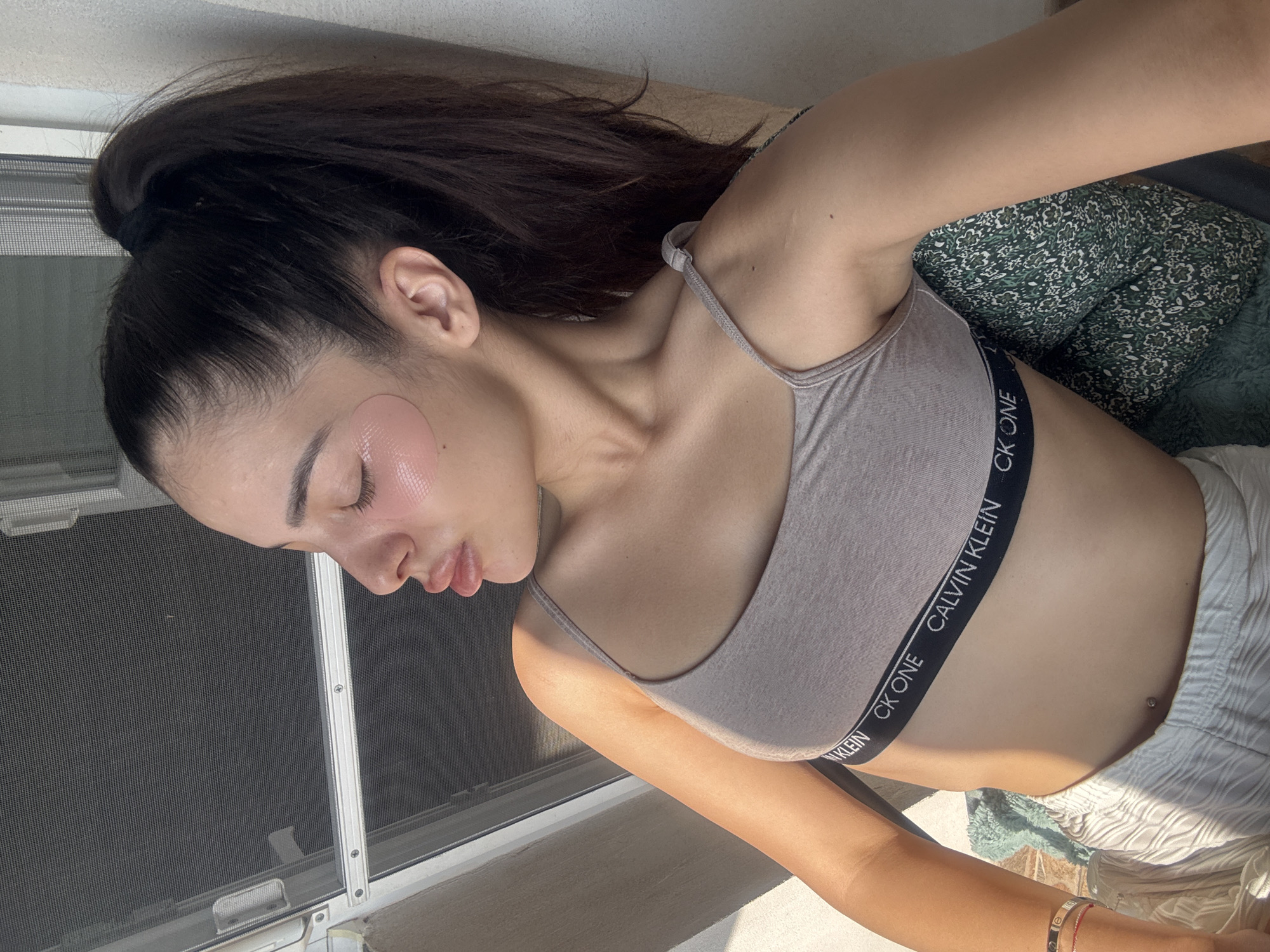 A young woman is depicted in a relaxed, sunlit pose, with her head tilted to the side and eyes closed or downcast. She has dark hair styled in a high ponytail. She wears a light beige bralette or sports bra with the text "CALVIN KLEIN CK ONE" visible on its elastic band. Her lower attire consists of light-colored, possibly white or cream, textured shorts. Two translucent, pinkish skincare patches are applied to her cheekbones.
Bright natural sunlight streams into the scene, creating strong highlights and shadows, indicating it is daytime. A window screen with metal framing is visible to her left, suggesting she is indoors near a window or on a balcony. A green and white patterned pillow supports her upper body, and a lighter-colored wall or surface is in the background. A small belly button piercing is visible on her midriff, and a bracelet adorns her right wrist. The overall impression is one of tranquility and self-care, possibly in Plovdiv, Bulgaria.
