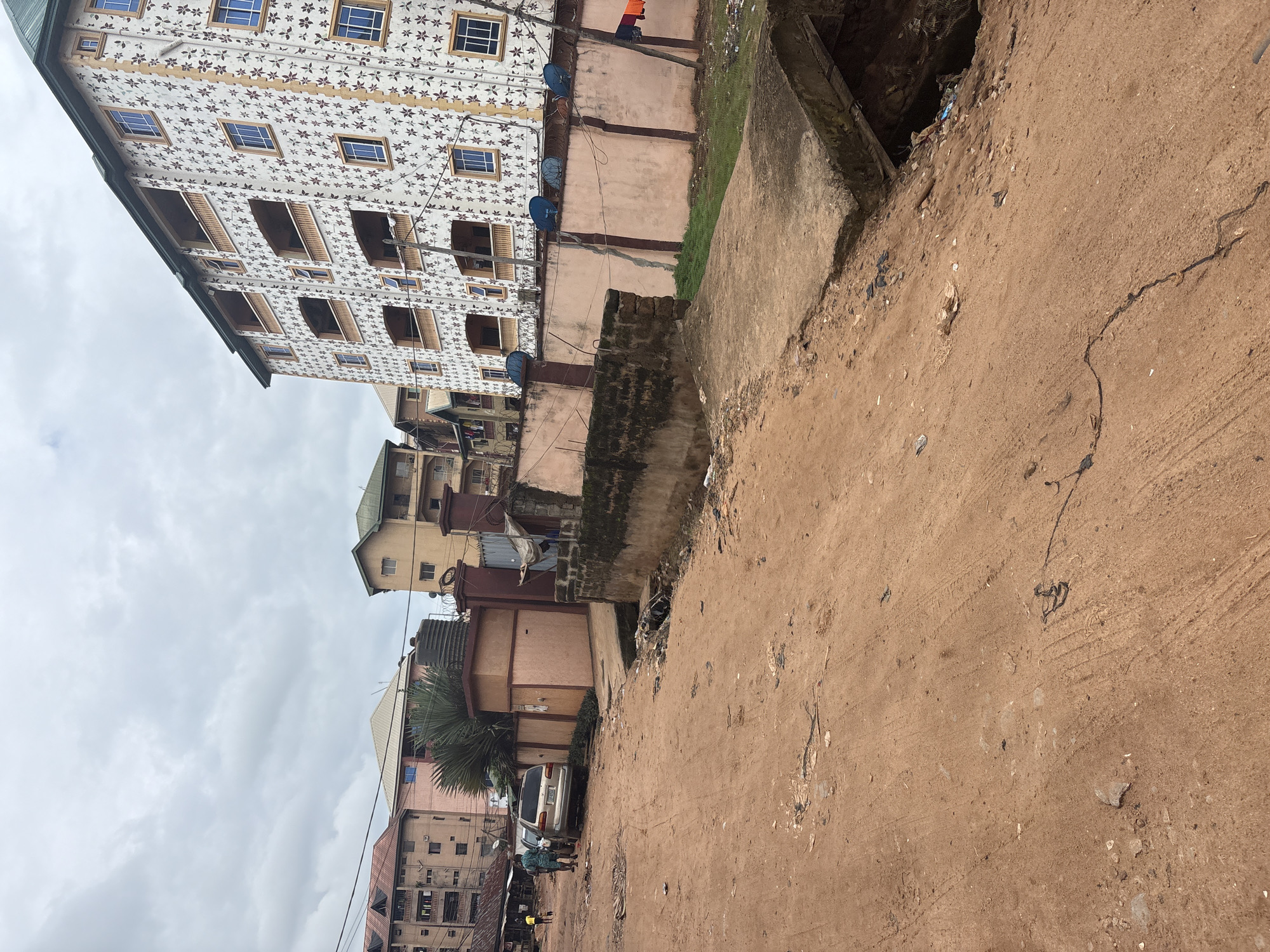This is a daytime, eye-level, wide shot of a dirt road in Nkpor, Nigeria. The road is unpaved and appears worn, with tire tracks visible. To the right, a concrete structure, possibly a bridge or culvert, spans an open drainage ditch that is filled with debris. Beyond the ditch is a pink wall with a patterned facade and several satellite dishes mounted on top. Behind the wall, a tall, multi-story building with a white exterior decorated with a floral pattern rises, featuring multiple balconies and blue-tinted windows. To the left of the dirt road, other multi-story buildings with similar architectural styles are visible, some with balconies. Two people are standing near a silver car parked on the side of the road. The sky is overcast with grey clouds. The scene depicts a common urban environment with infrastructure, residential buildings, and signs of daily life in Nkpor.