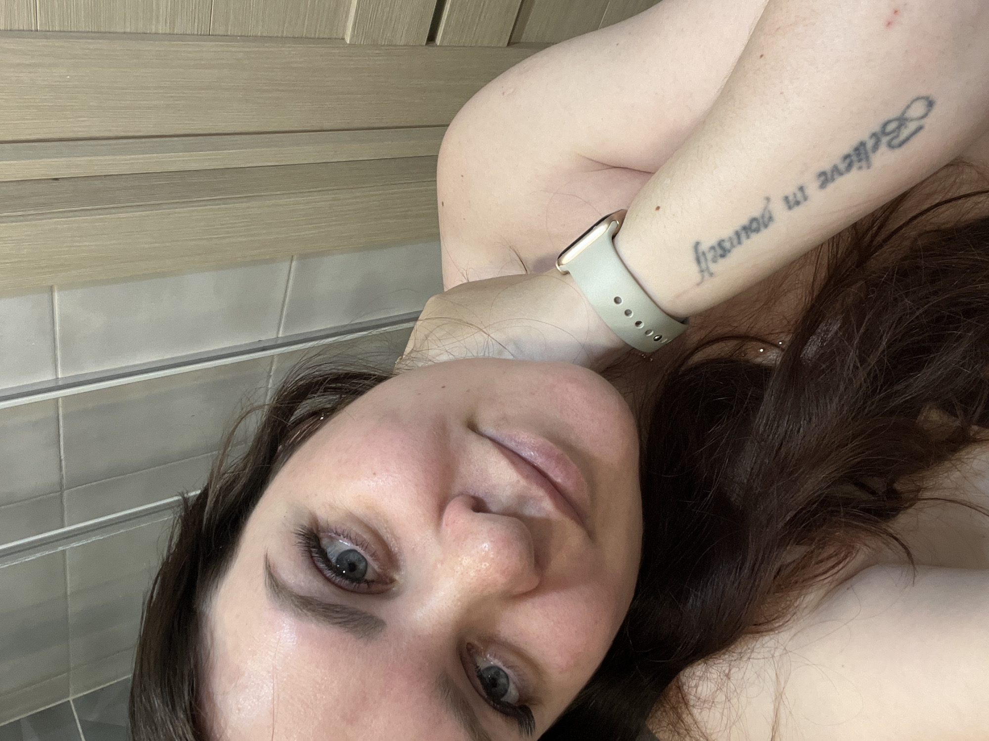 A close-up, slightly angled selfie of a fair-skinned woman with long, dark brown hair. Her eyes are light-colored and appear to be blue or gray. She is wearing a light-colored smartwatch on her left wrist, and a tattoo reading "Believe in yourself" is visible on her inner forearm. The background includes light-colored wood paneling above gray tiles. The photograph was taken indoors. There is no discernible indication that the location is Noviy, Russia.