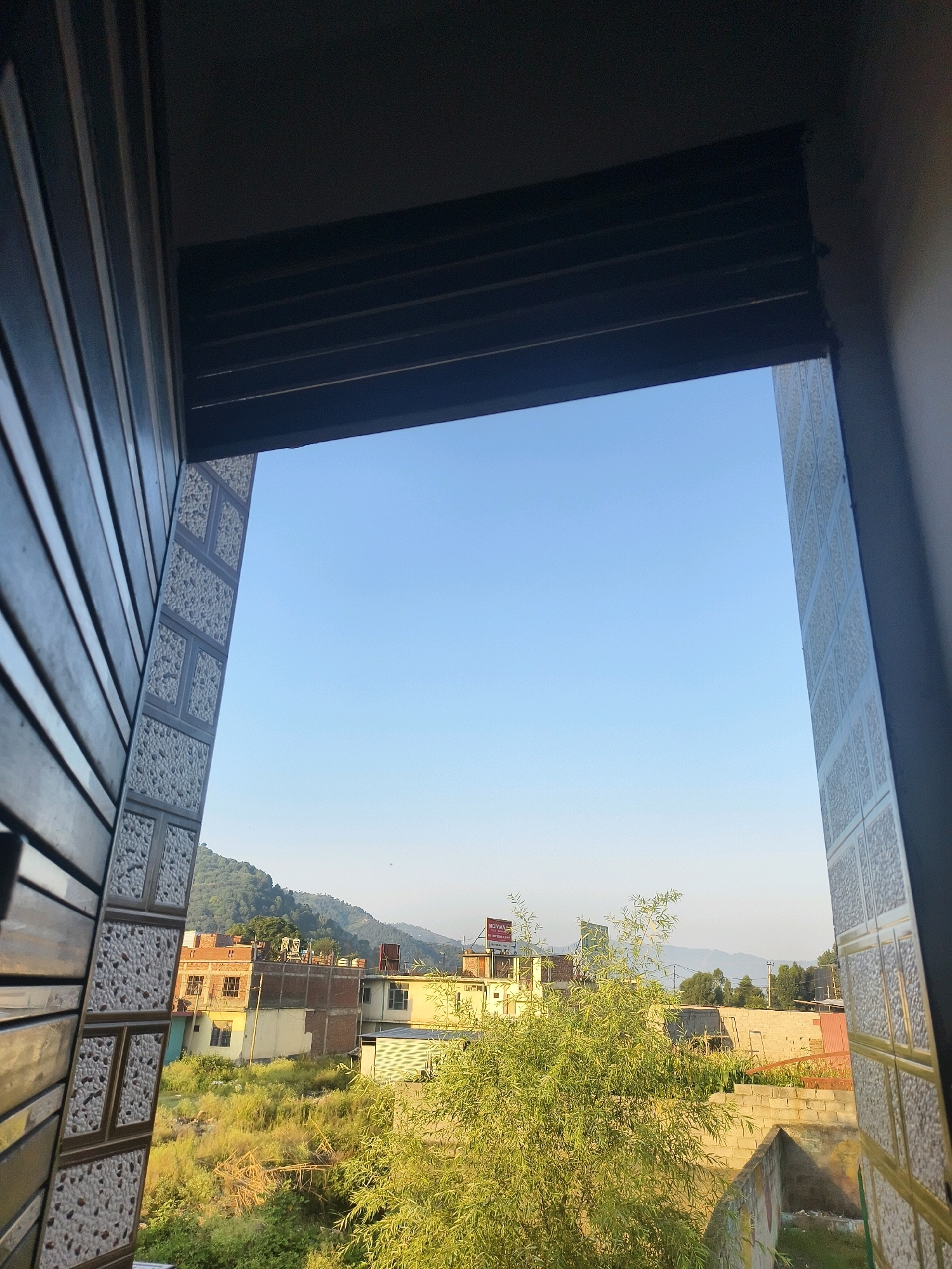The image captures a view through an open window or doorway in Poonch, India, looking out onto a landscape with buildings, vegetation, and hills under a clear blue sky. In the foreground, on the left side, is a portion of a building's exterior with a textured pattern and vertical slats. On the right side, a similar textured pattern is visible on a wall. The middle ground features several buildings, some appearing to be residential or commercial, with brick and concrete construction. There are also areas of dense green vegetation, including trees and shrubs. In the background, rolling green hills are visible, suggesting a semi-urban or rural setting on the outskirts of the city. The lighting suggests it is daytime, likely morning or late afternoon given the soft light. There are no people or animals clearly visible in the frame. The scene depicts a common view from a dwelling overlooking a developing area with natural elements.