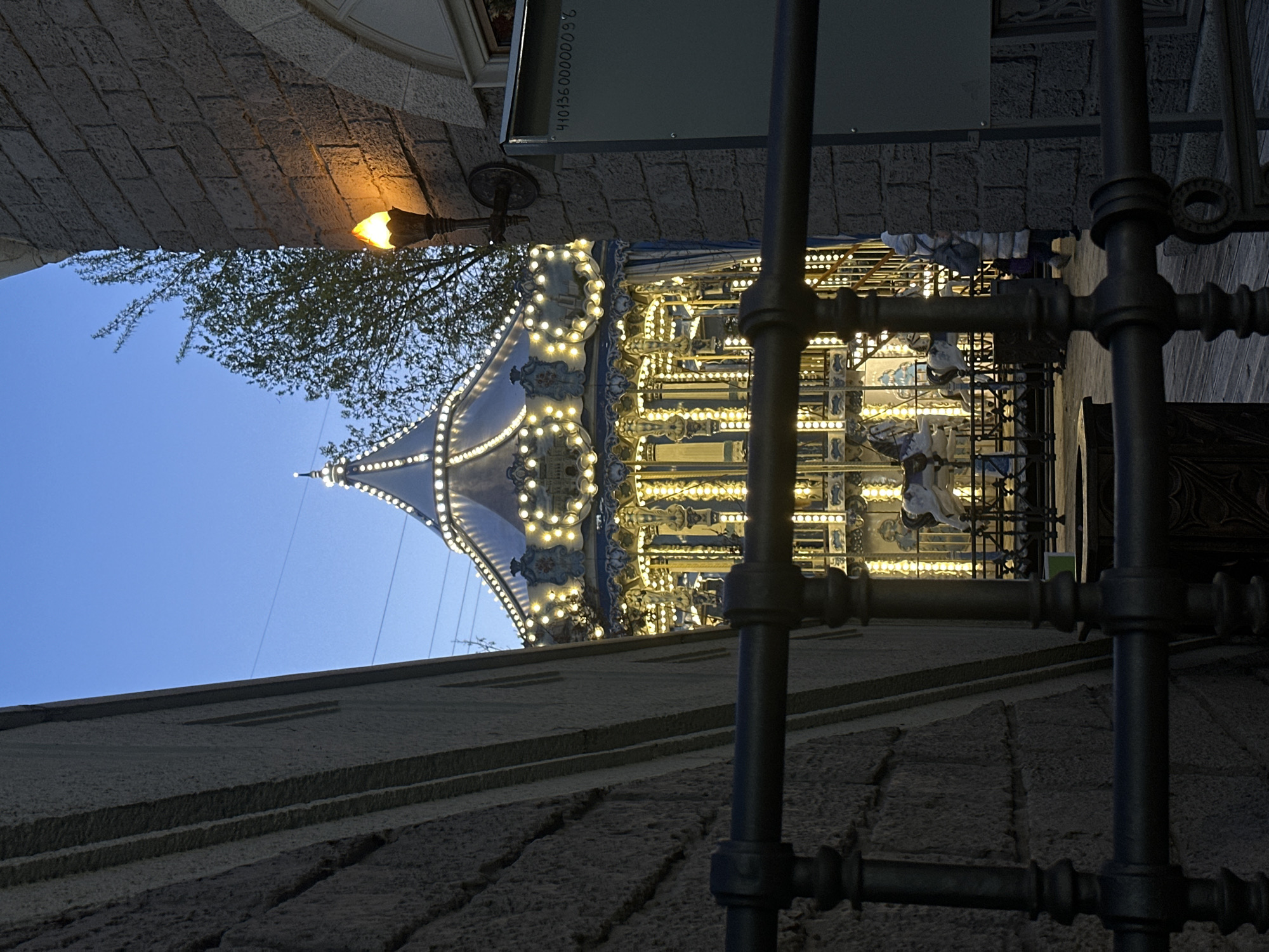 Captured from a low angle, the photograph features a carousel silhouetted against a twilight sky, framed by architectural elements of Moscow, Russia. The carousel, illuminated with strings of lights, showcases a conical canopy and moving figures, presumably horses, and is the focal point. A portion of a stone building is visible in the foreground and background, presenting layered stonework. Metal scaffolding or piping runs vertically in the foreground. A small, antique-style lamp is attached to the stonework, casting a warm glow on the wall. Above the wall, the sky transitions from a light blue to a darker hue, suggesting the onset of dusk. A tree with sparse foliage stretches into the sky alongside the carousel's canopy. The overall scene exudes a whimsical yet historic atmosphere, characteristic of Moscow's urban landscape.
