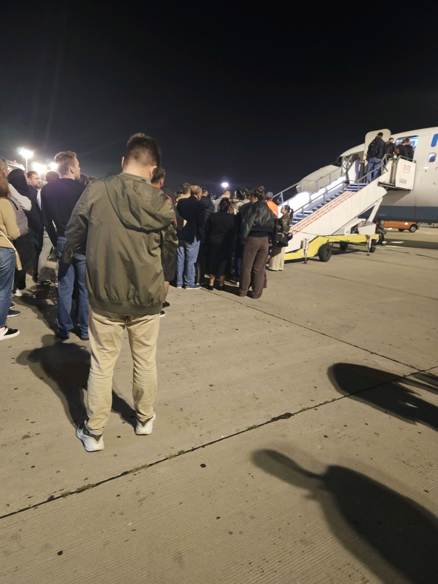 The image depicts a nighttime scene at an airport tarmac, specifically at Vnukovo, Russia. A group of passengers are queuing to board an airplane via an airstair. The passengers are varied in age and attire, with most wearing casual clothing. The airstair is illuminated, and the airplane itself is visible, showing a portion of its fuselage with airline branding. Shadows of the people and the airstair are cast on the concrete tarmac, indicating artificial lighting. The background is dark, suggesting it is night. Some ground equipment, including what appears to be a baggage cart, is also present.