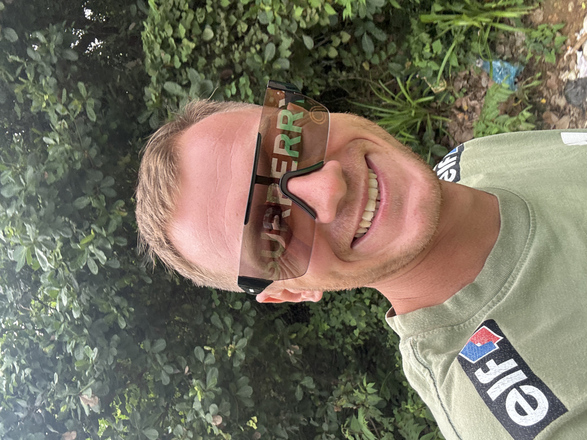 A man is taking a selfie in front of a leafy green background. He is wearing a light green t-shirt with an "Elf" logo on the left shoulder and large, reflective sunglasses with the word "BURBERRY" visible on the lenses. He has short, blond hair and is smiling, showing his teeth. The background appears to be a dense area of foliage, possibly a garden or jungle, with some discarded items visible on the ground in the lower right portion of the image. The lighting suggests it is daytime. The location is identified as Rawai, Thailand.