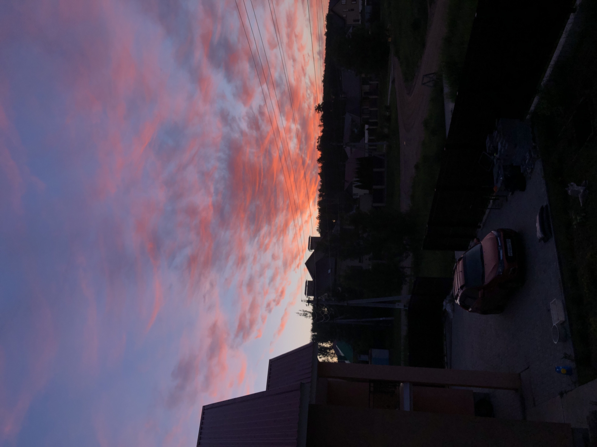 The image depicts a serene sunset over a residential area in Noviy, Russia, viewed from an elevated perspective, possibly from a building's upper floor or balcony. The sky is a dominant feature, filled with dramatic, streaky clouds painted in vibrant hues of orange and pink, blending into purples and soft blues towards the upper left.
Along the horizon, a dark silhouette of tree lines and various house rooftops is visible against the colorful sky. Several parallel power lines stretch across the mid-ground, cutting through the vibrant clouds. Below the horizon line, numerous houses are nestled among trees, appearing dark and shadowed in the fading light.
In the foreground on the lower left, part of a building's brown roof and structure are visible. On the lower right, a red car, likely a sedan or hatchback, is parked on a paved driveway or yard. A blurry license plate can be seen on its rear. Next to the car, there's a dark, indistinct object, possibly covered. A dirt path or unpaved road is also faintly visible winding through the background on the right.
The scene captures the transition from day to night, characterized by the striking colors of the sunset and the darkening landscape. No people are visible, and there is no specific activity taking place, conveying a sense of quietude and natural beauty at dusk.