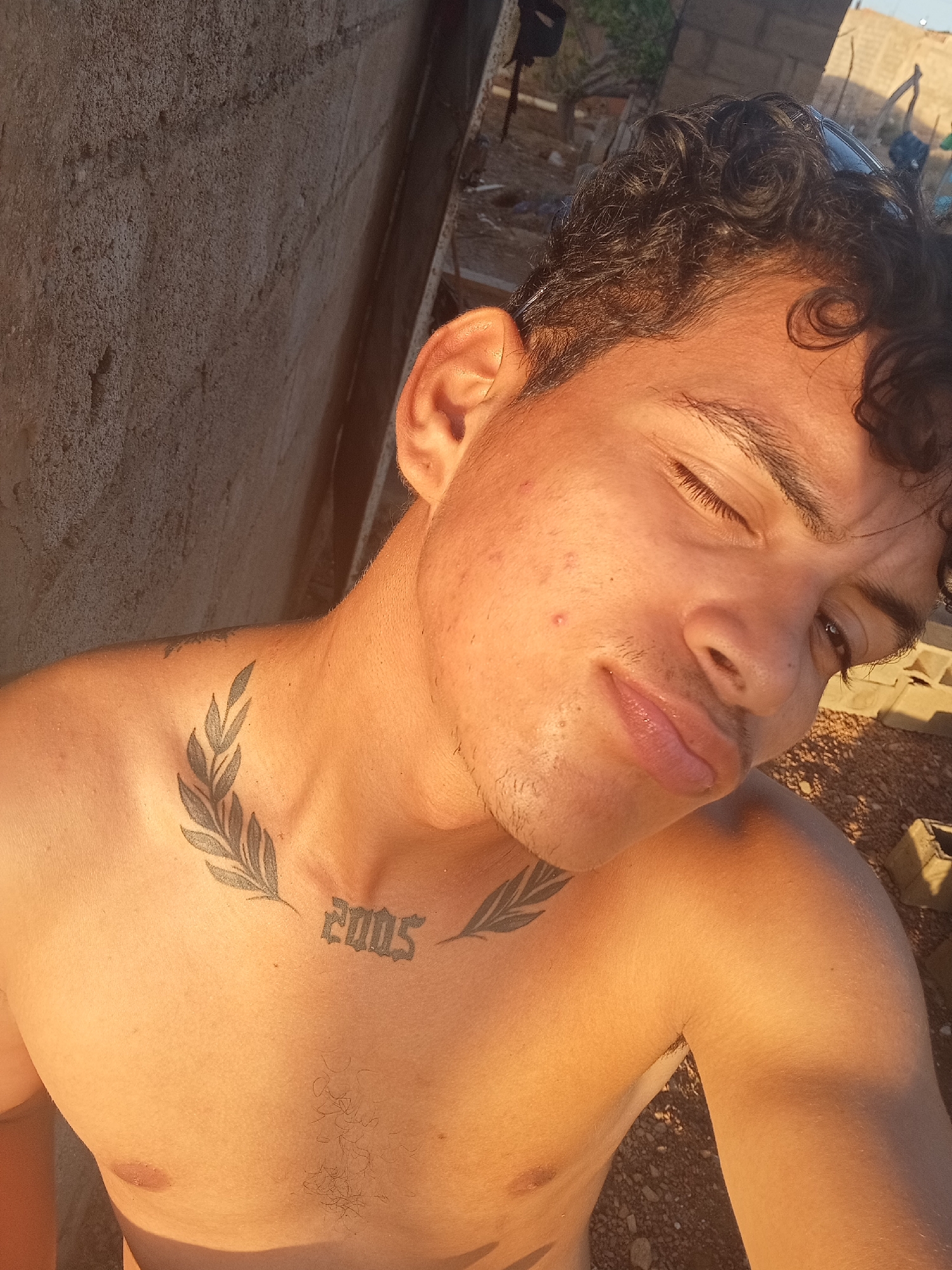 A young, shirtless man with dark, curly hair is posing for a selfie. He is fair-skinned, with a slightly unshaven face. The man is looking at the camera with one eye partially closed. He has a tattoo on his chest featuring foliage and the number "2005". The background is sunlit, with a block wall visible on the left and what appears to be construction debris and sparse vegetation visible to the right and rear. Sunglasses are perched on the top of the man's head. Based on the provided information, the location is Altagracia, Venezuela, though there are no explicit indications of the city in the photo.