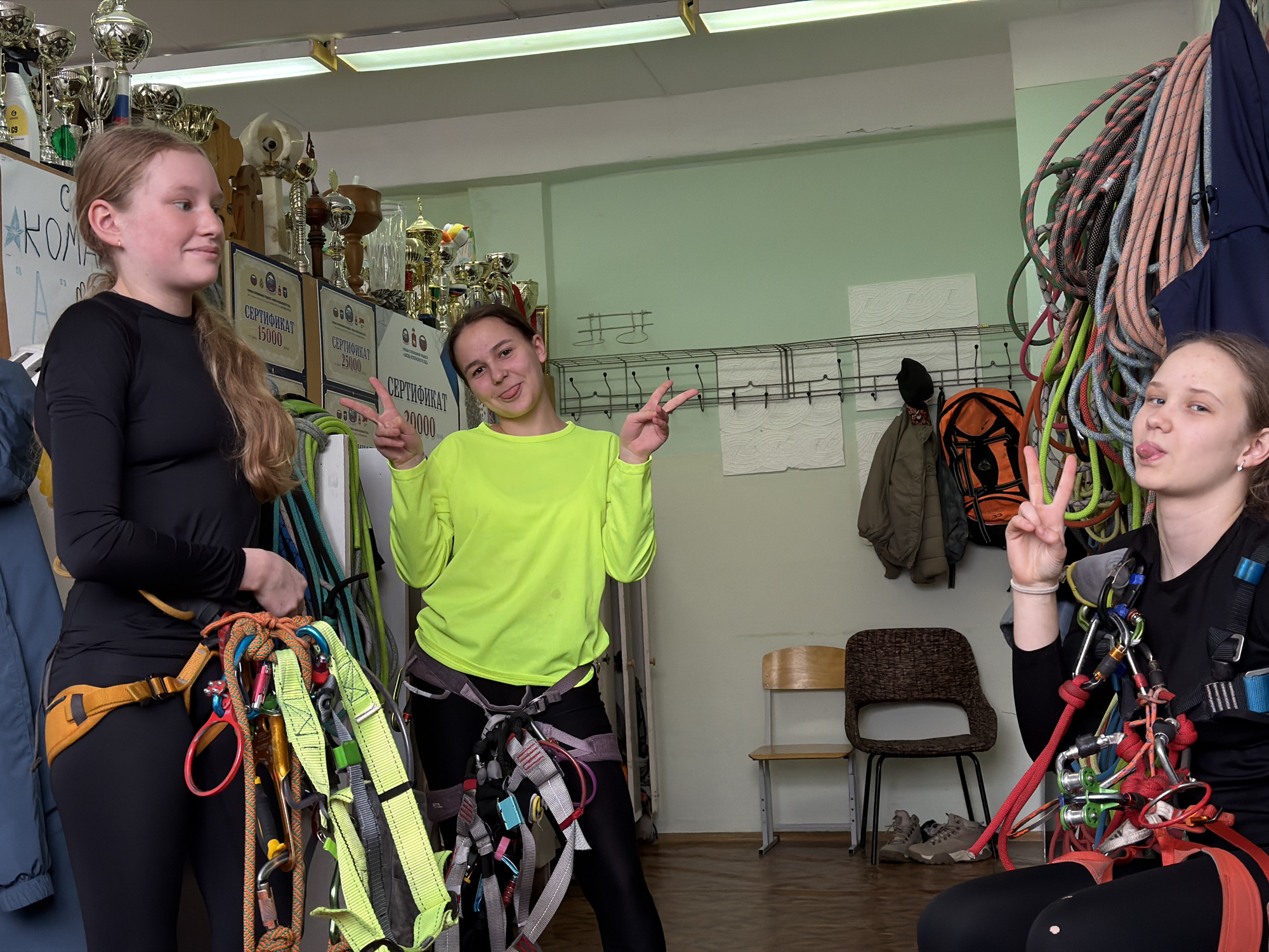 Three young women are posing indoors. They are all wearing climbing harnesses with various pieces of climbing equipment attached. The woman on the left is wearing a black long-sleeved shirt and black leggings. The woman in the middle is wearing a bright yellow long-sleeved shirt and black leggings. The woman on the right is wearing a black long-sleeved shirt and black leggings and is sticking out her tongue while holding up a peace sign. The background includes climbing ropes, a rack with hanging items like a coat and a backpack, and two chairs. Trophies are visible on a shelf in the background on the left, along with certificates. This image was taken at the climbing wall in Berezniki, Russia.